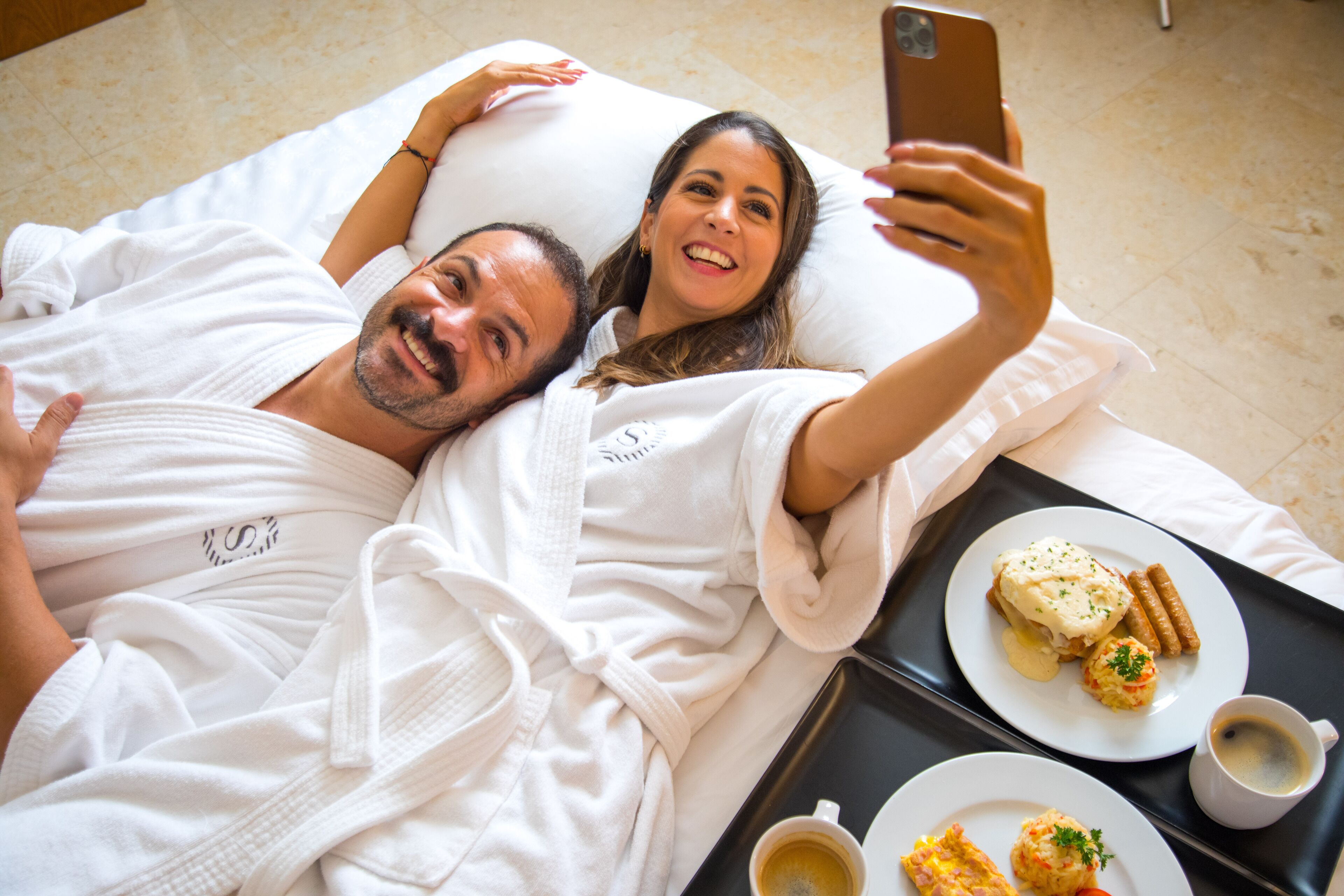 A couple lying in bed, taking a selfie, with breakfast including eggs, sausages, and coffee on a tray.