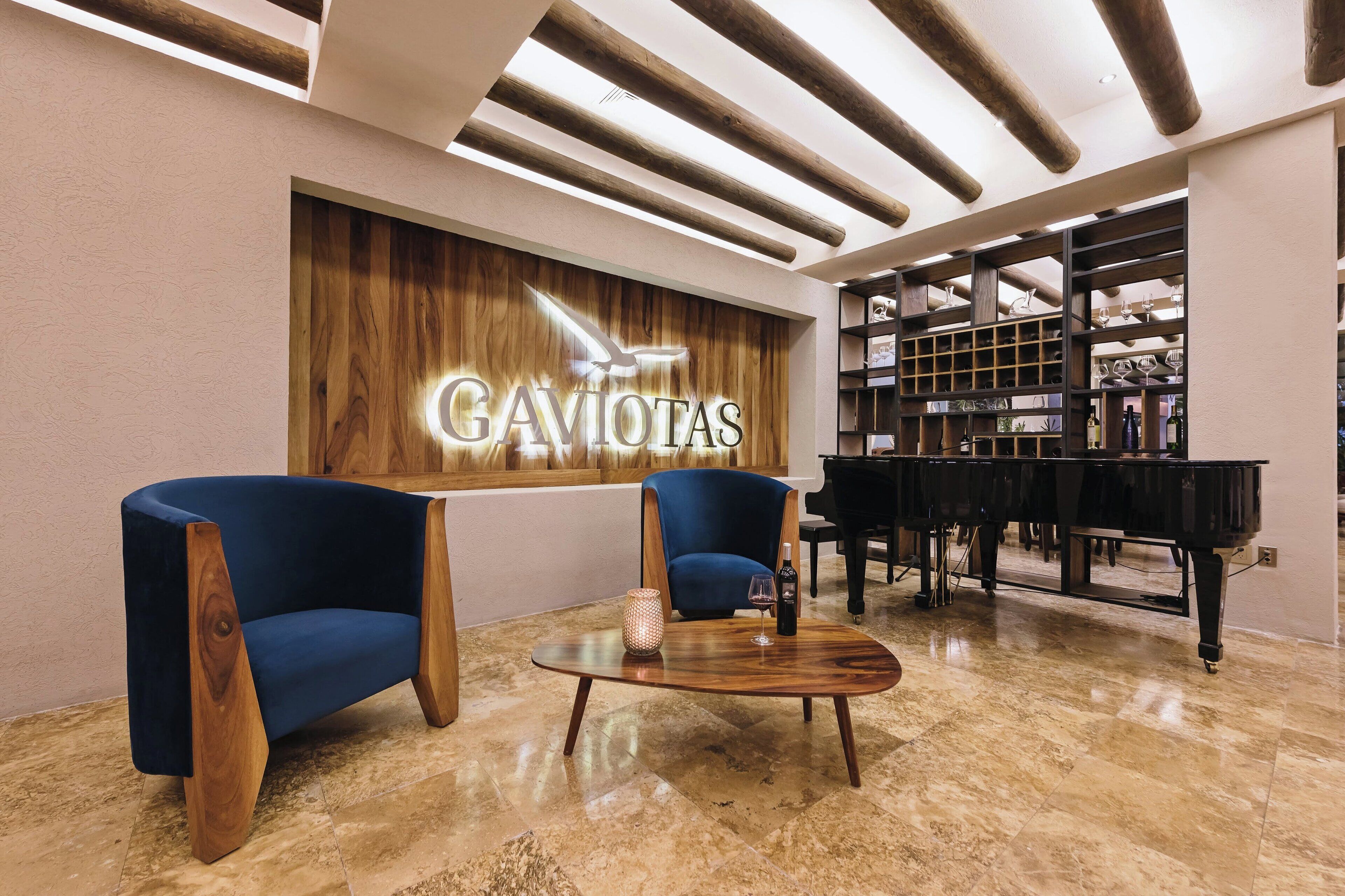 A lobby with two blue armchairs, a wooden coffee table, a piano, and a wall with 'GAVOTAS' signage.
