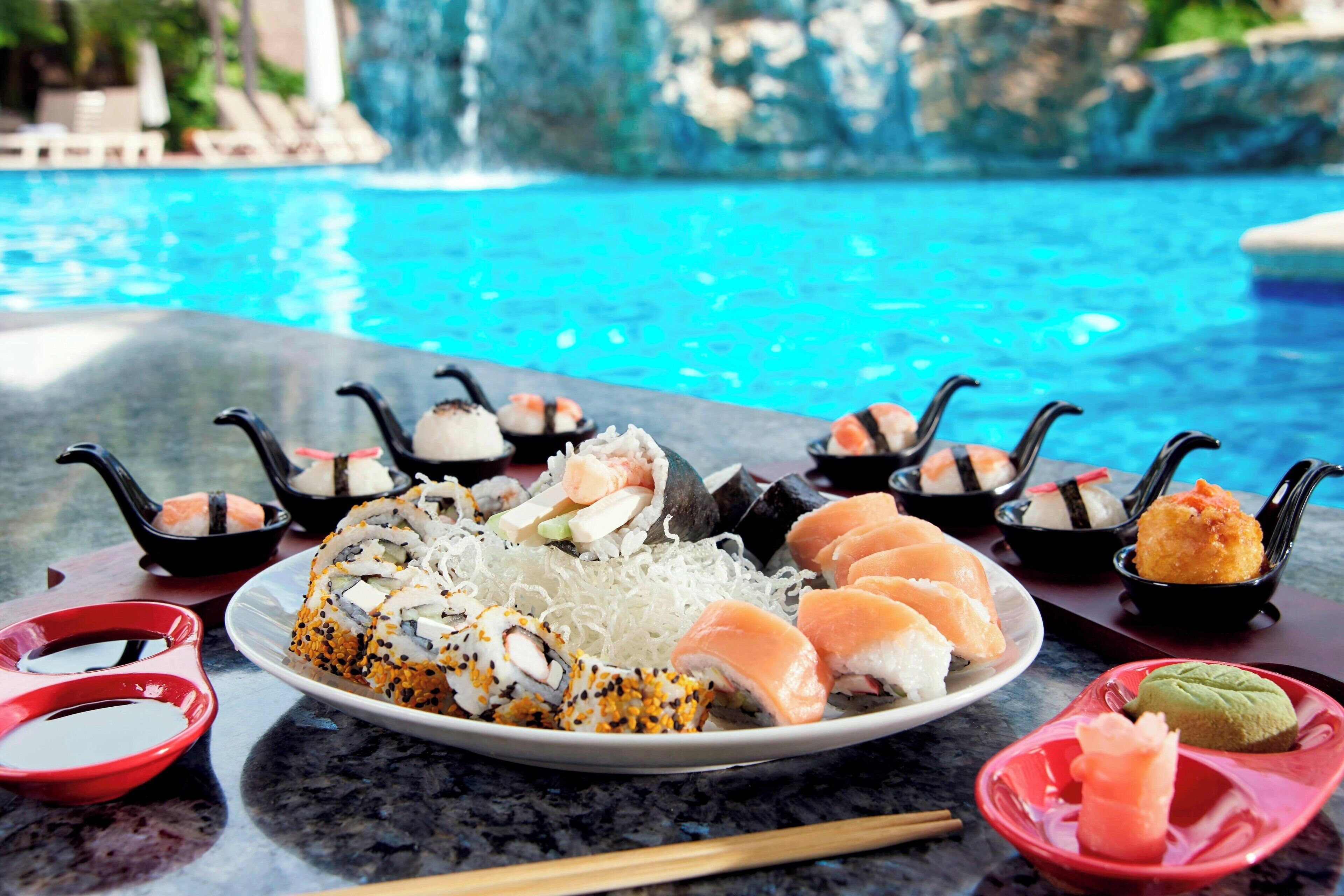 Un plateau de sushis comprenant différents types de sushis et de sashimis, accompagné de petits bols de sauce soja, le tout présenté sur fond de piscine et de paysage rocheux.