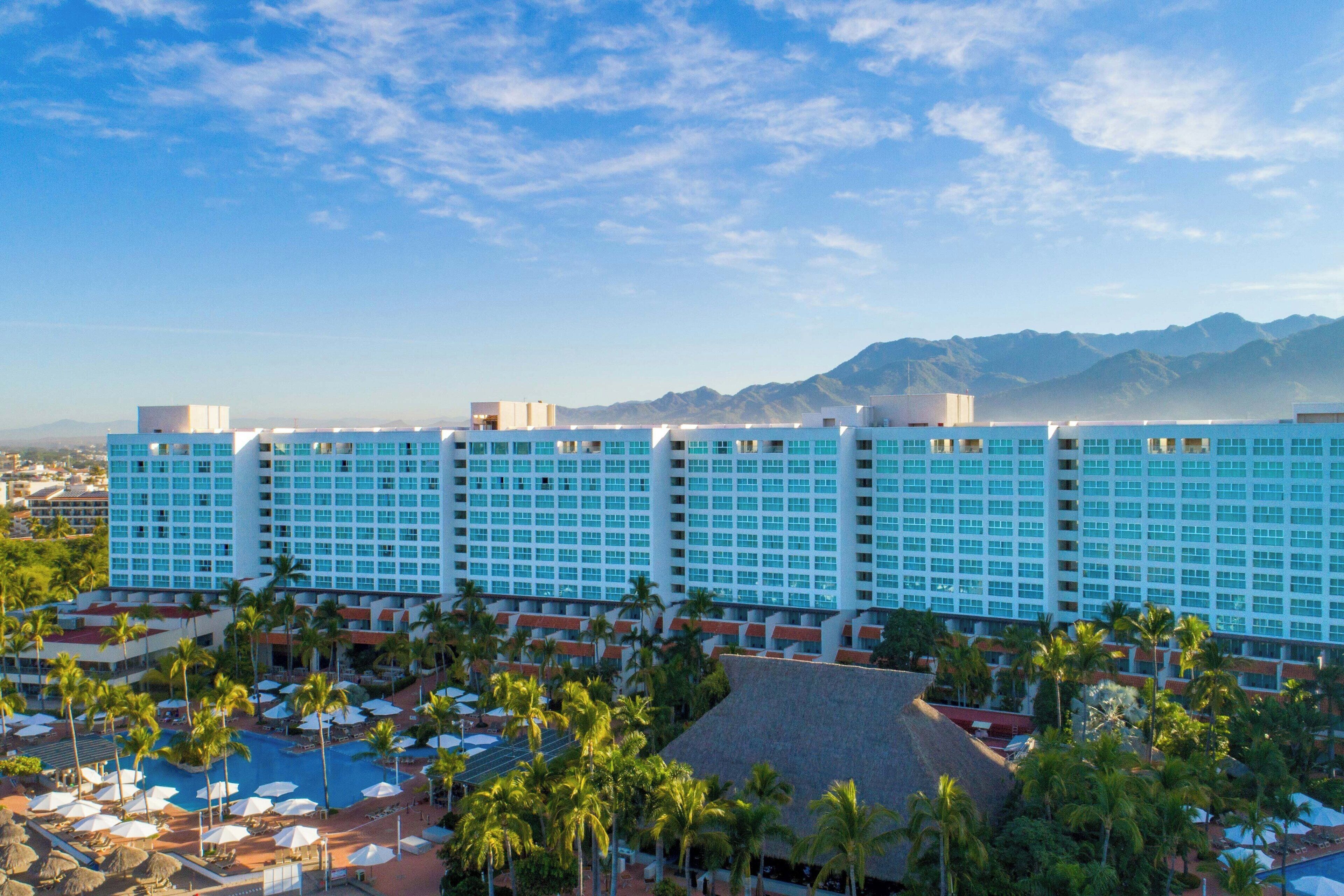 A hotel with a large number of rooms, a swimming pool, and palm trees.