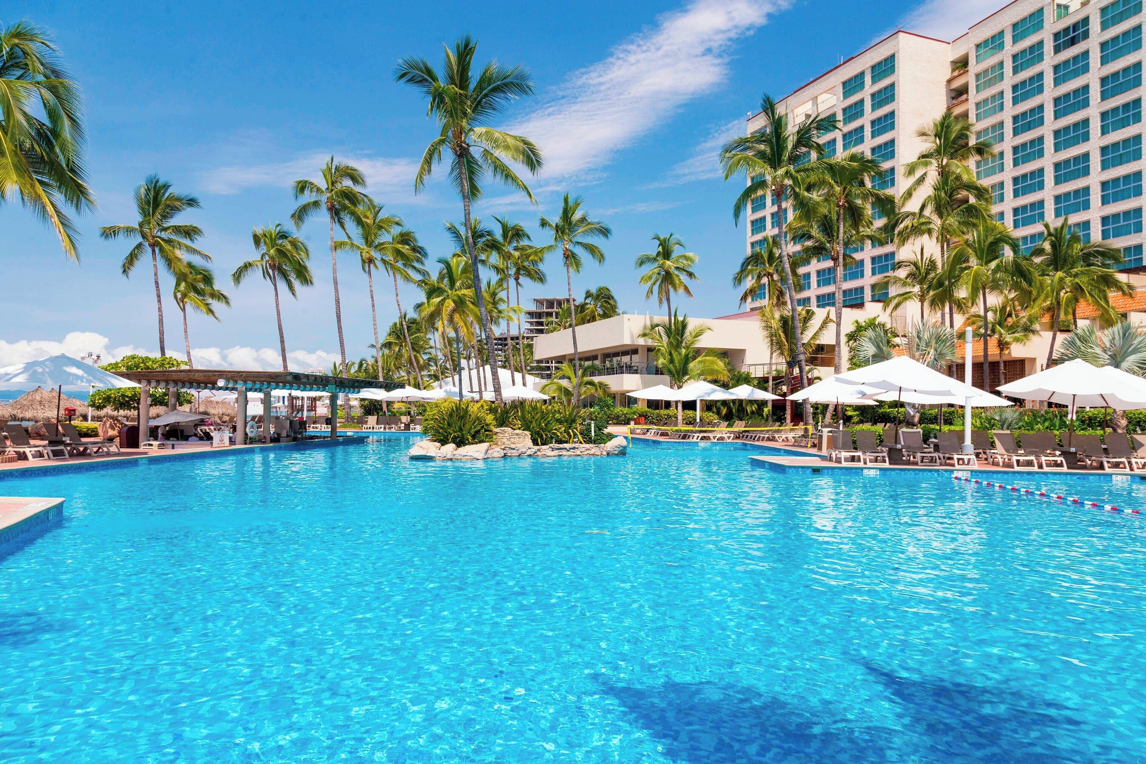 A large outdoor swimming pool surrounded by palm trees and lounge chairs, with a hotel building in the background.