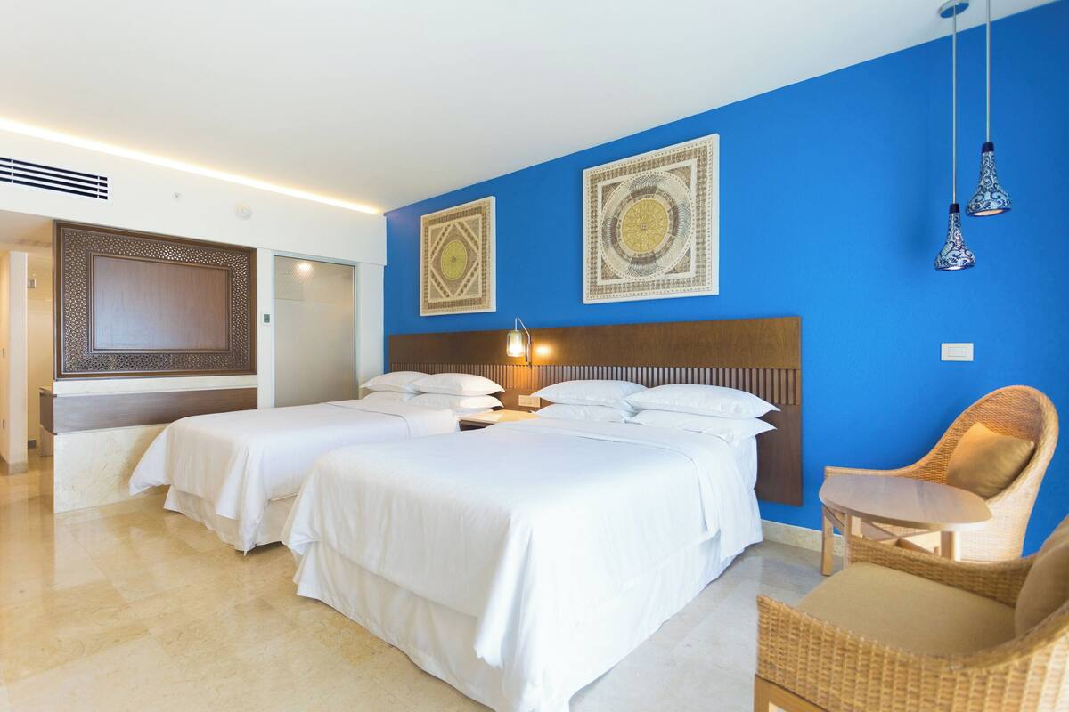 Ein Hotelzimmer mit zwei Betten, einem hölzernen Kopfteil, einem Korbstuhl und Wanddekoration.