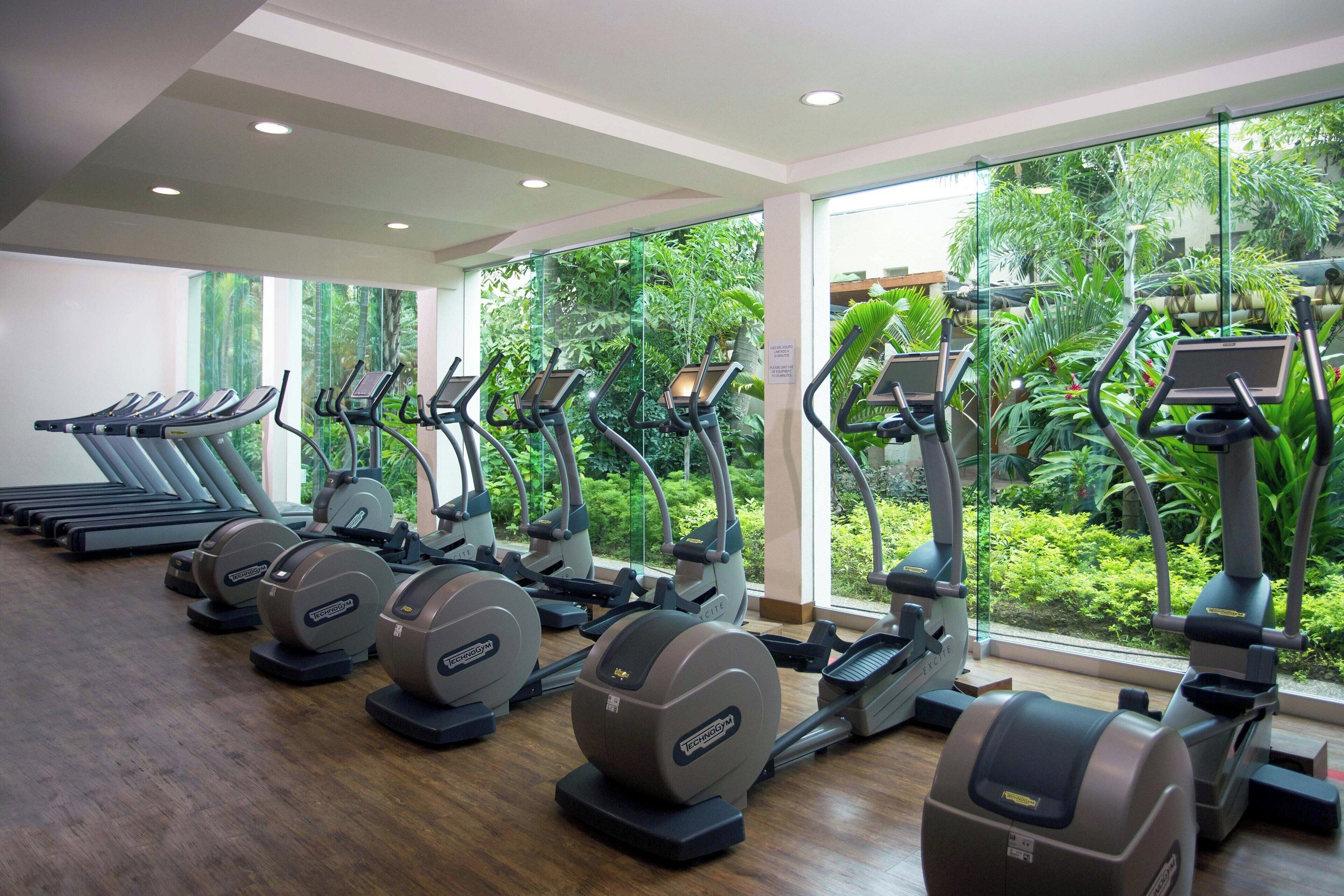 A gym with multiple treadmills and elliptical machines, large windows with a view of greenery.