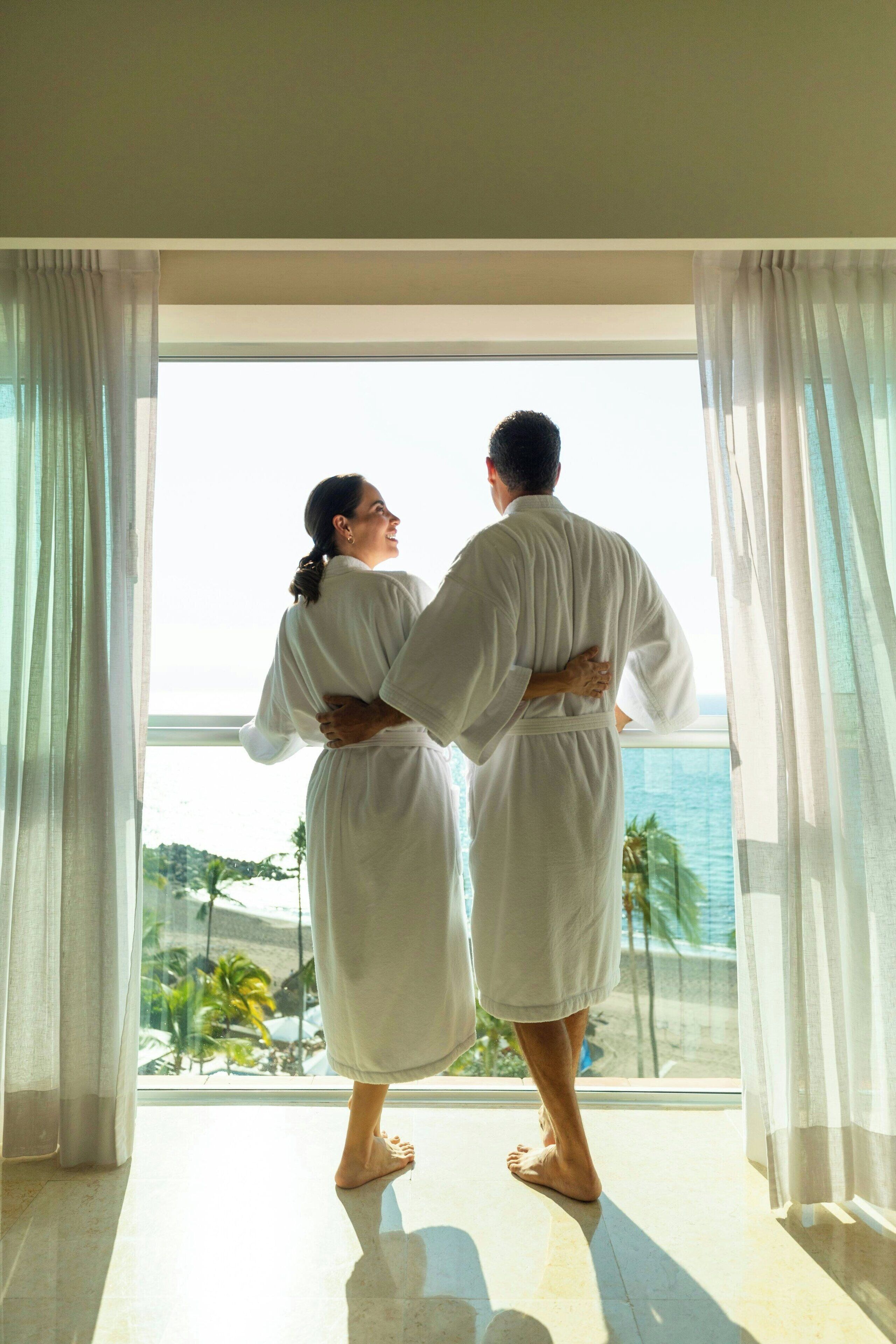 A couple standing by a large window with sheer curtains, overlooking a scenic view of the ocean and palm trees.