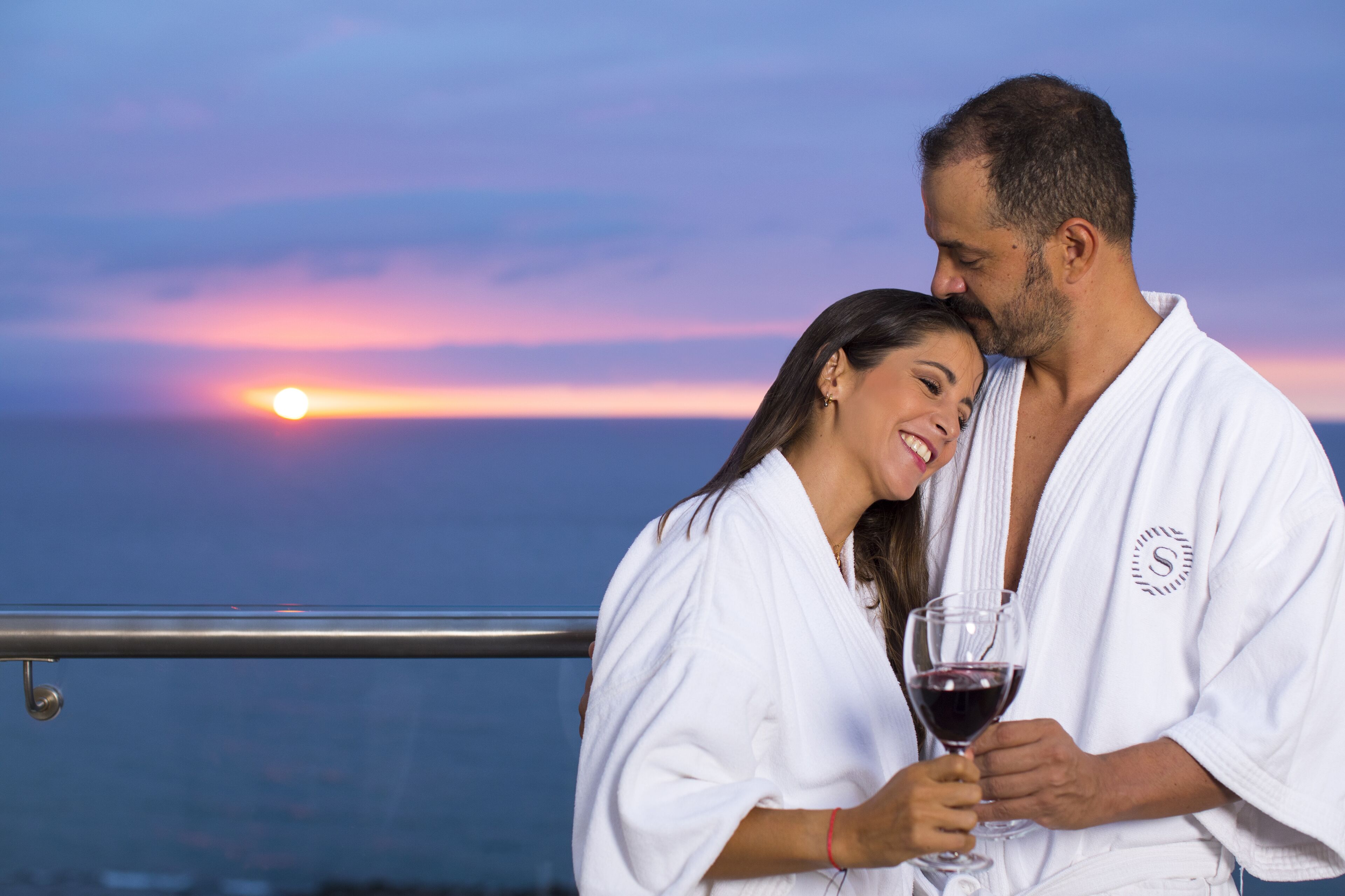 Una pareja vestida de blanco junto a una barandilla, con vista al atardecer.