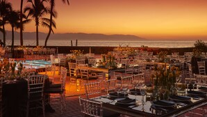 An outdoor dining area with tables and chairs, palm trees, and a view of the ocean at sunset. - Sheraton Buganvilias Puerto Vallarta  (Puerto Vallarta)