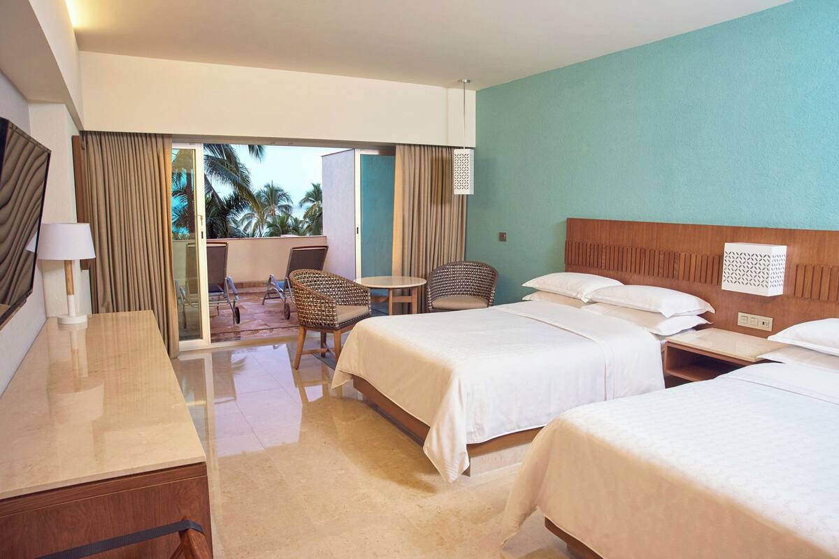 Ein Hotelzimmer mit zwei Betten, einem Schreibtisch, einem Stuhl und einem Balkon mit Blick auf Palmen.