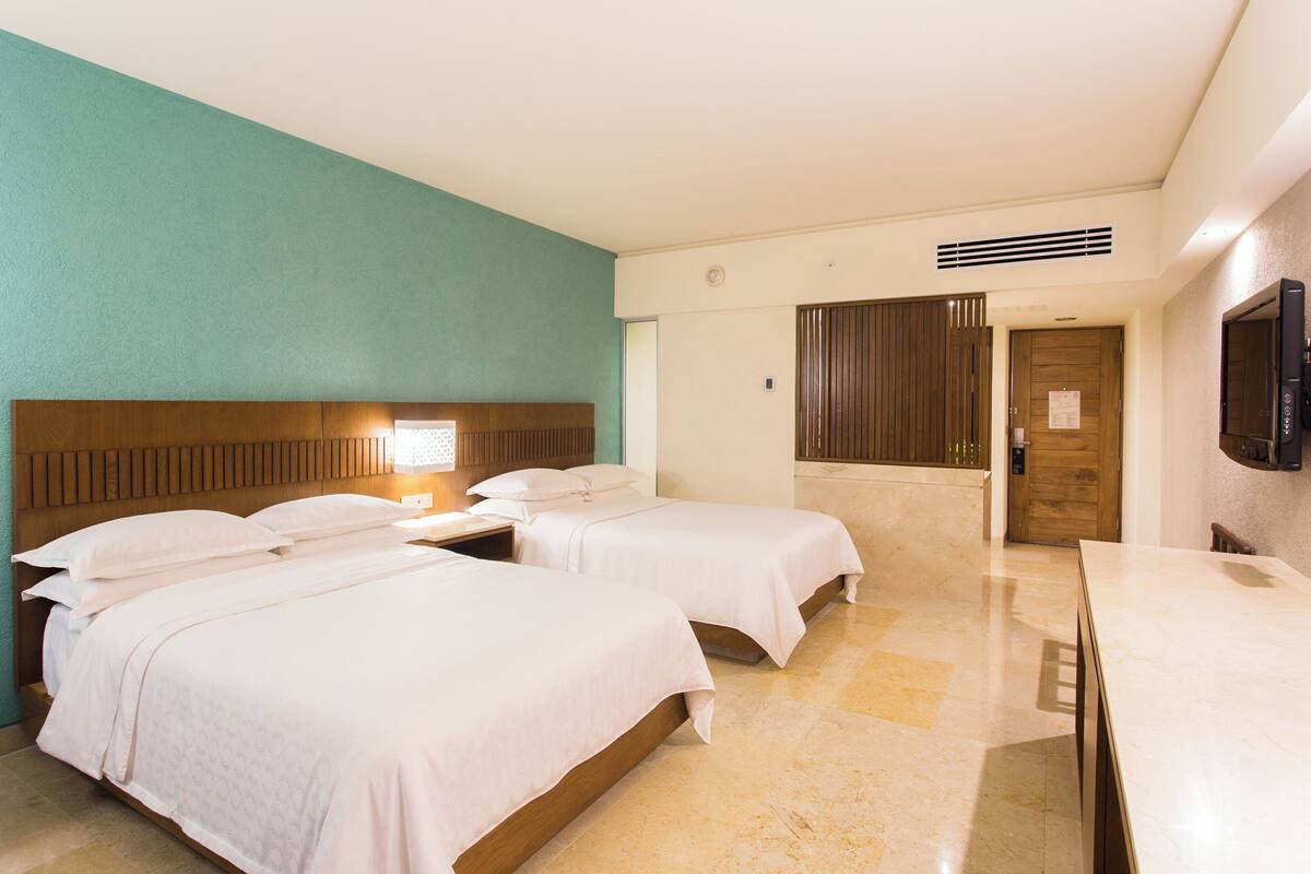 Ein Hotelzimmer mit zwei Betten, einem hölzernen Kopfteil, einem Flachbildfernseher und einem Badezimmer mit Dusche und Toilette.