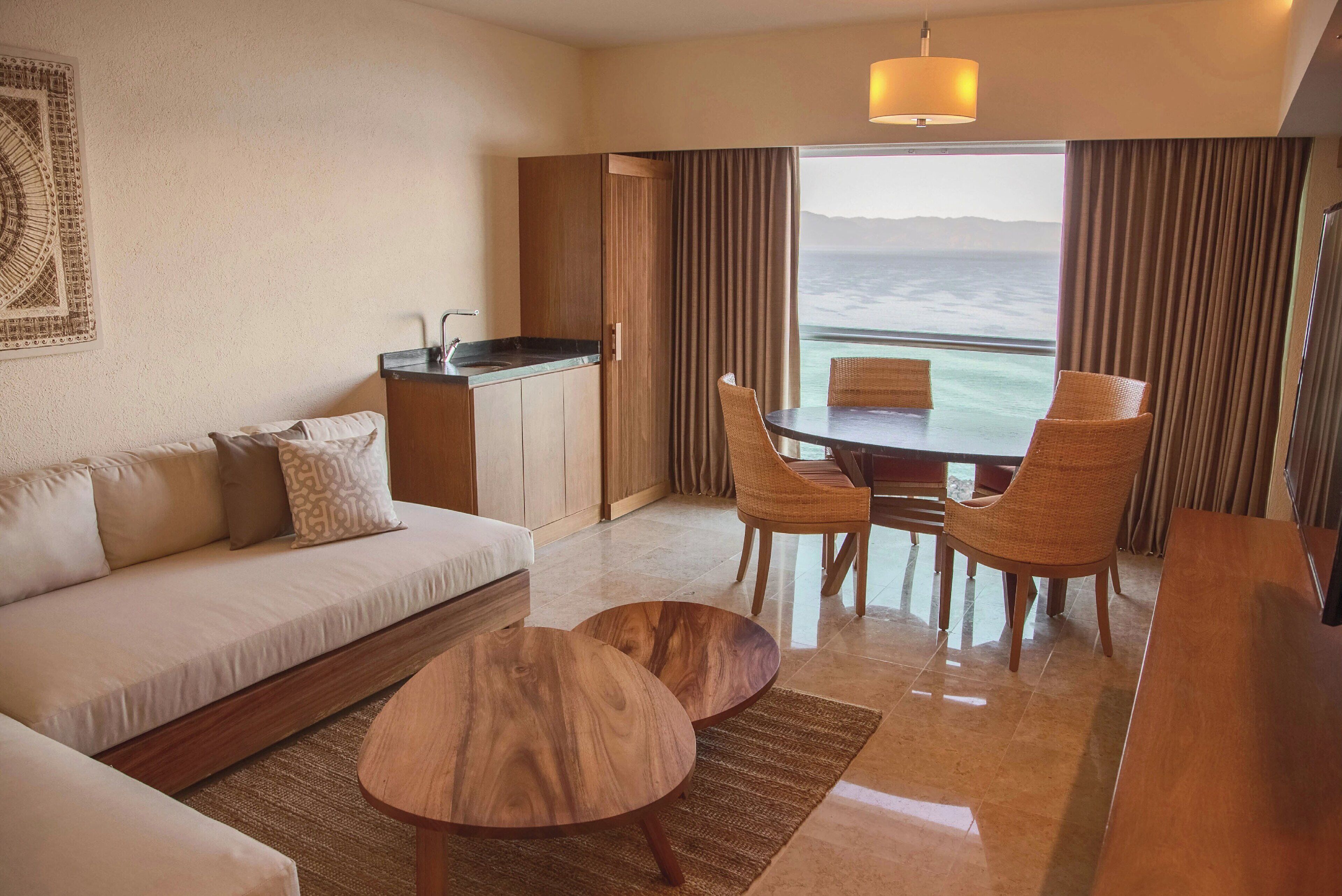 Habitación de hotel con sofá, dos mesas de centro de madera, una mesa de comedor con sillas, un área de cocina y vistas al océano.