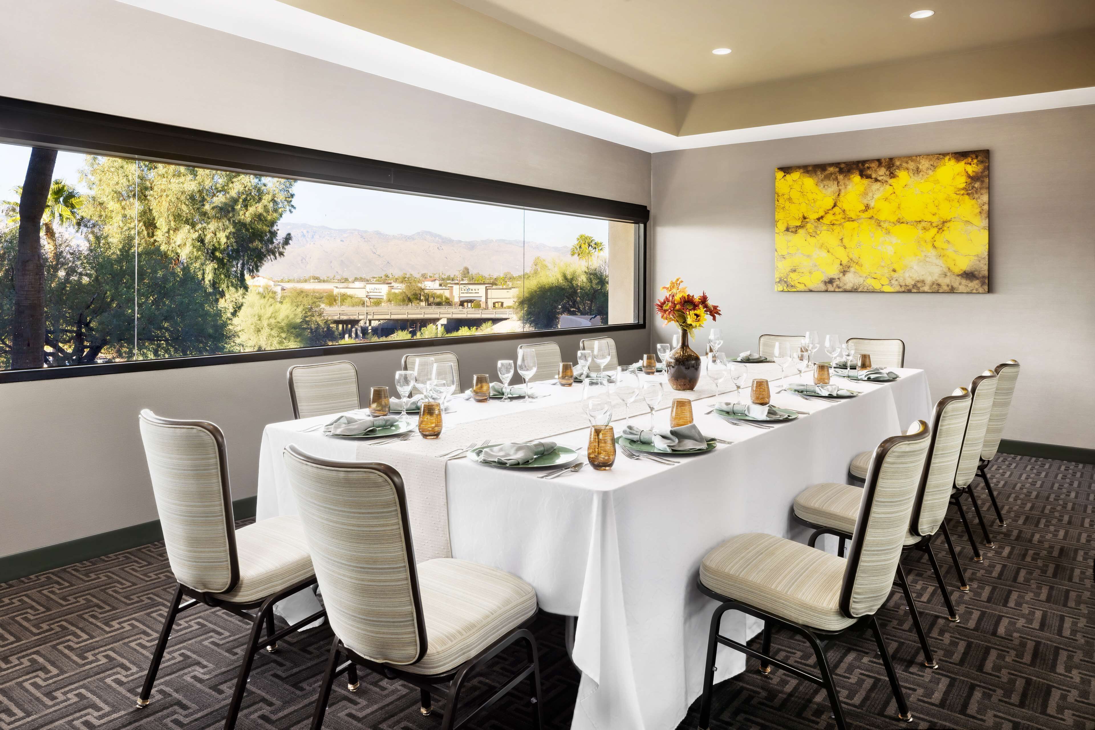 A conference room with a long table set for a meeting, chairs around the table, a window with a view of trees and mountains, and a yellow abstract painting on the wall.