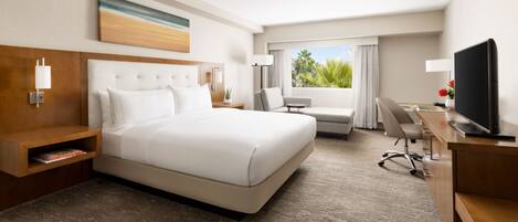 A hotel room with a large bed, a desk, a chair, a TV, and a window with a view of greenery.