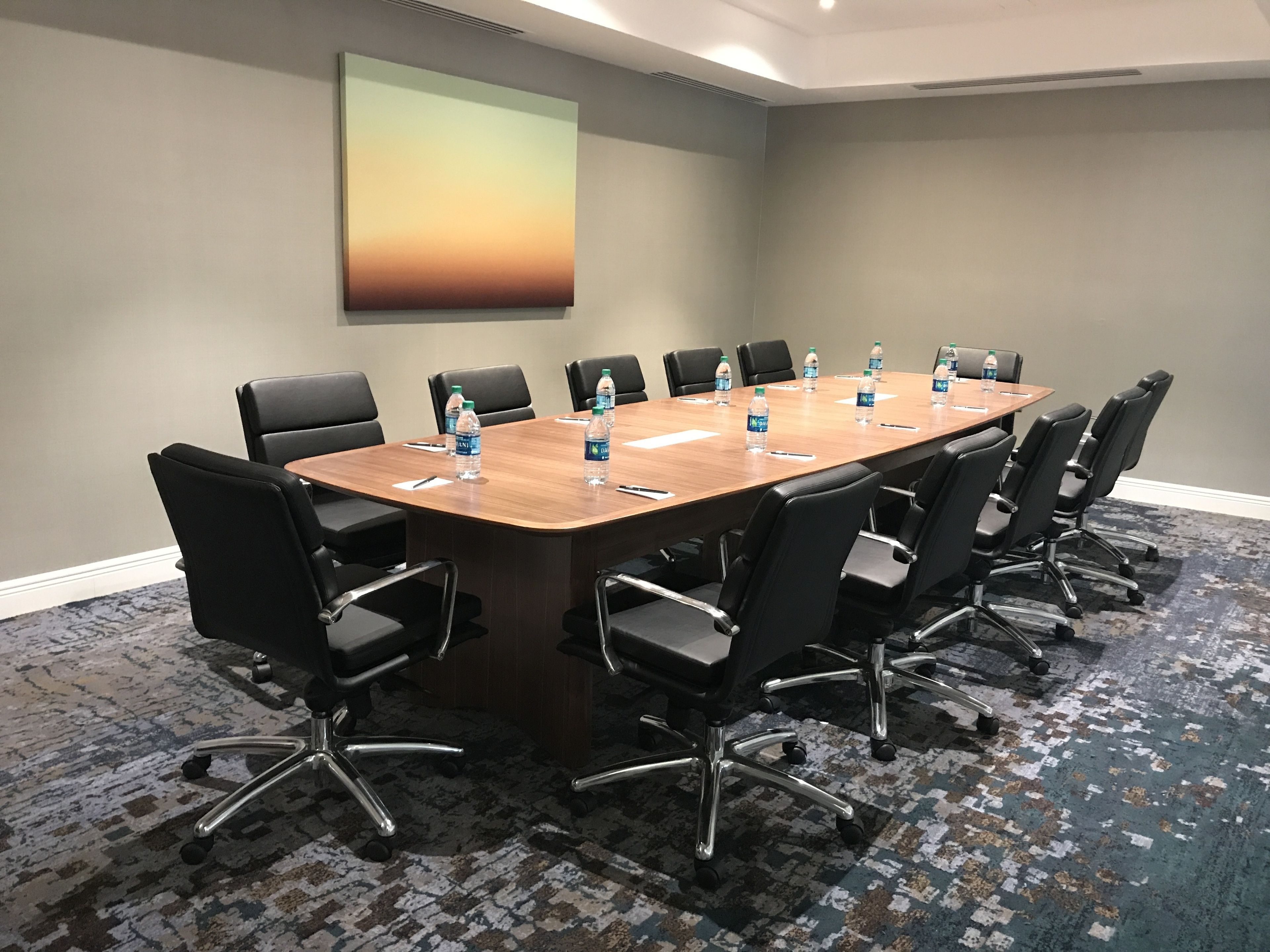 Un salón de conferencias con una larga mesa, sillas negras, botellas de agua y un cuadro abstracto grande en la pared.