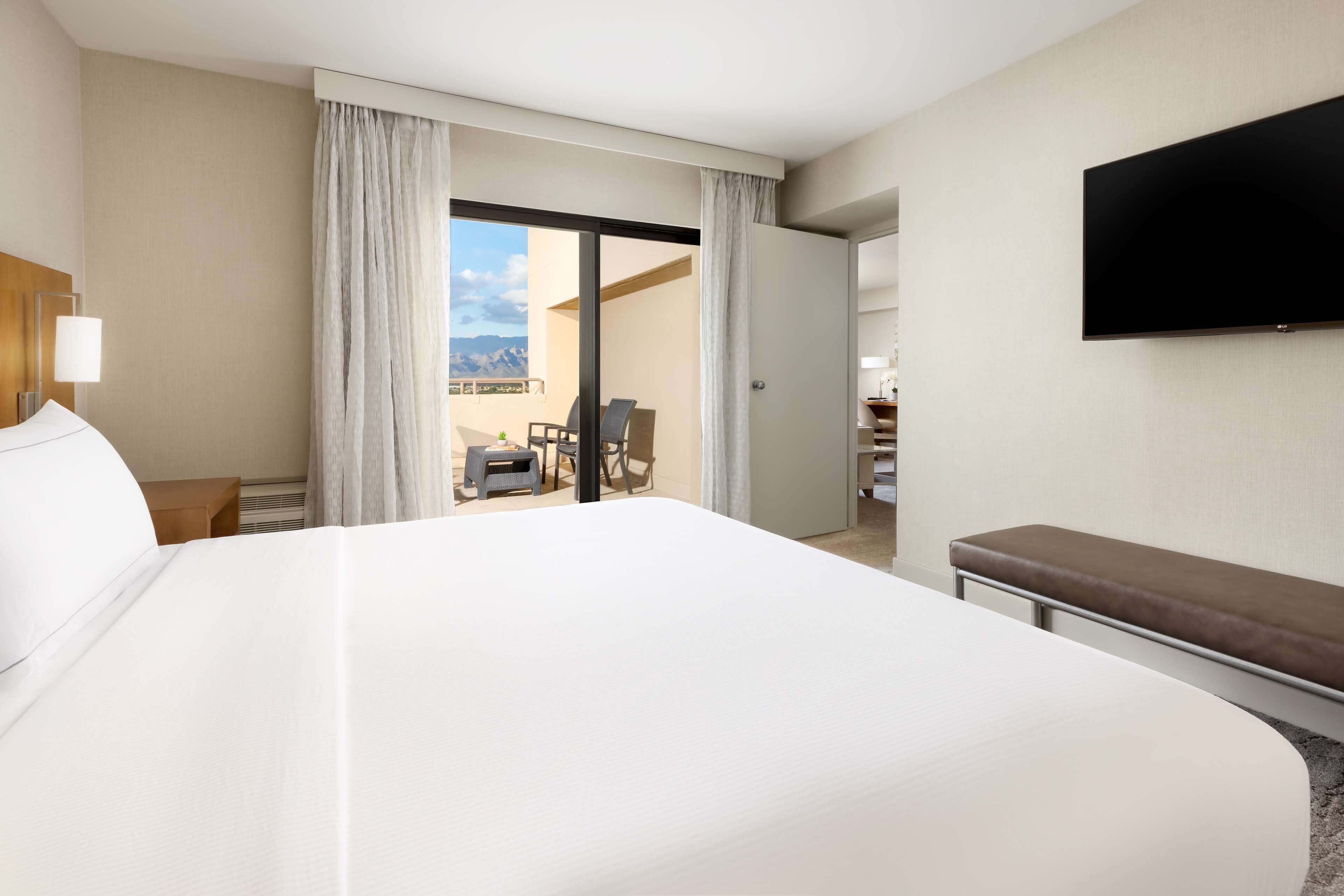 A hotel room with a large bed, a flat-screen TV mounted on the wall, a balcony with a chair and table, and a sliding door leading to a balcony with a view of the mountains.