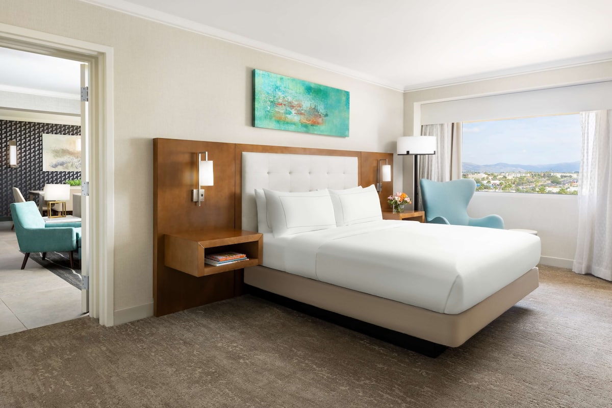 Quarto de hotel com uma cama grande, cabeceira de madeira, mesa de cabeceira com abajur, vista da cidade através de grandes janelas e um quadro na parede.