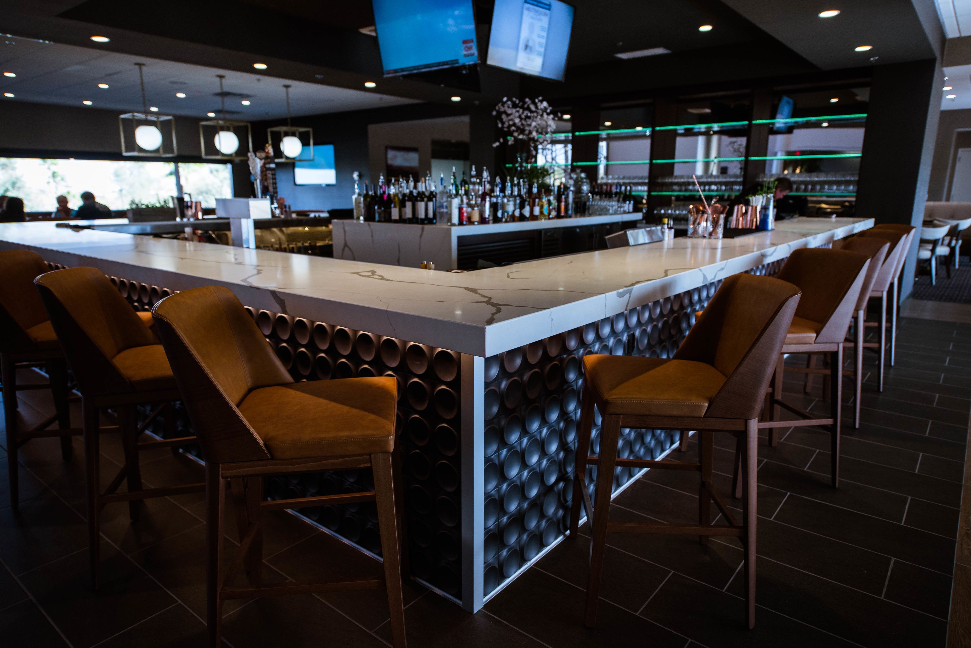 Zona de bar con mostrador de mármol, taburetes acolchados y una variedad de botellas y copas.