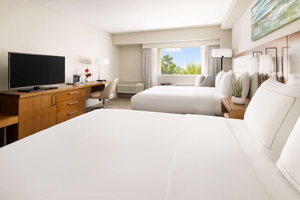 A hotel room with a large bed, a desk with a flat-screen TV, a chair, a desk lamp, and a window with a view of palm trees.