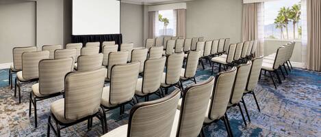 A conference room with rows of chairs, a projector screen, and large windows with curtains.