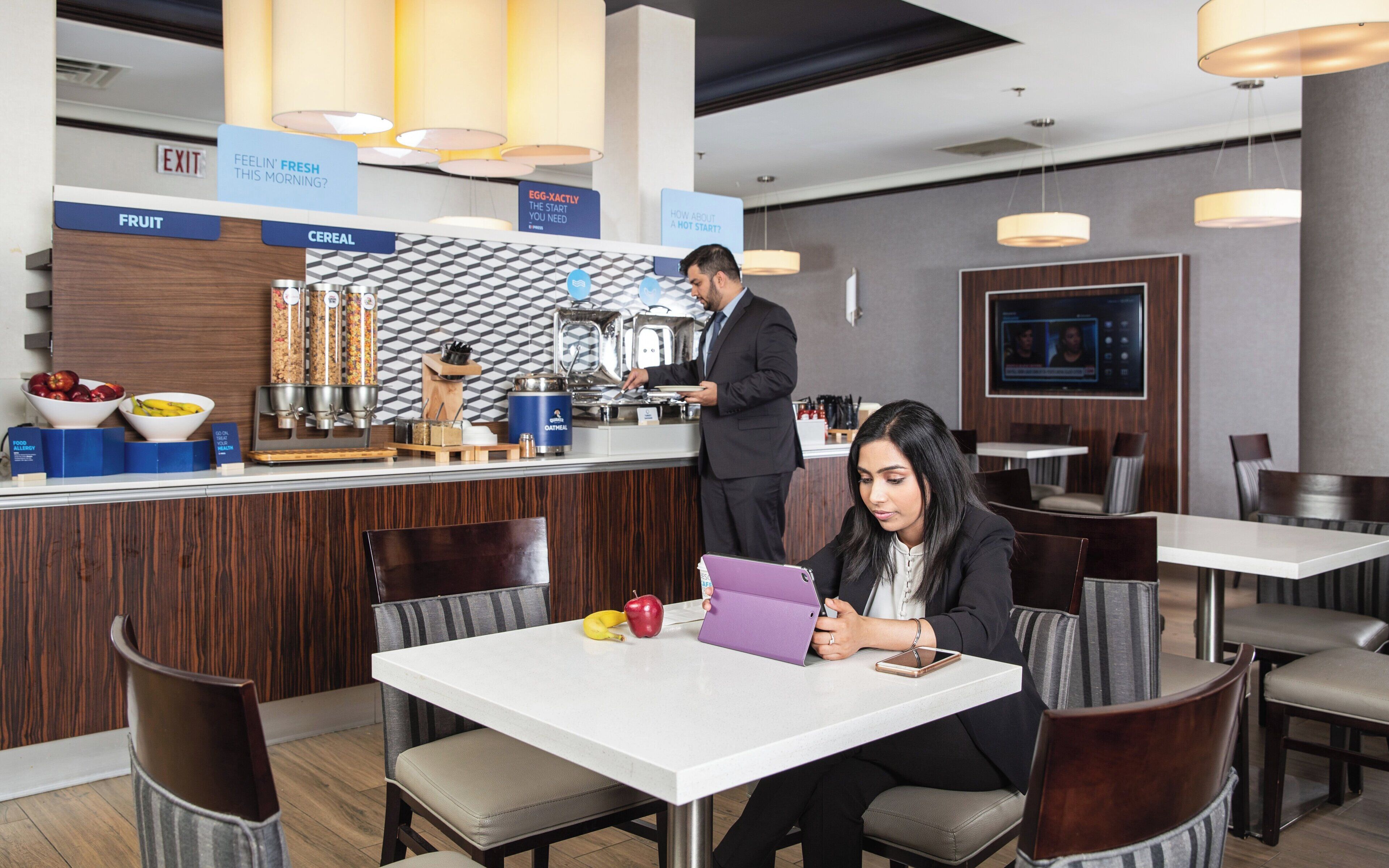 A hotel restaurant with a counter, a man serving food, and a woman using a tablet at a table.