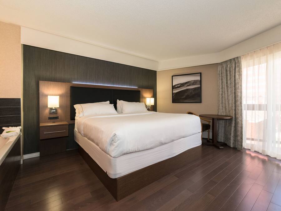 Quarto de hotel moderno com uma cama grande, mesas de cabeceira com luminárias, uma pequena escrivaninha e uma janela ampla com cortinas.