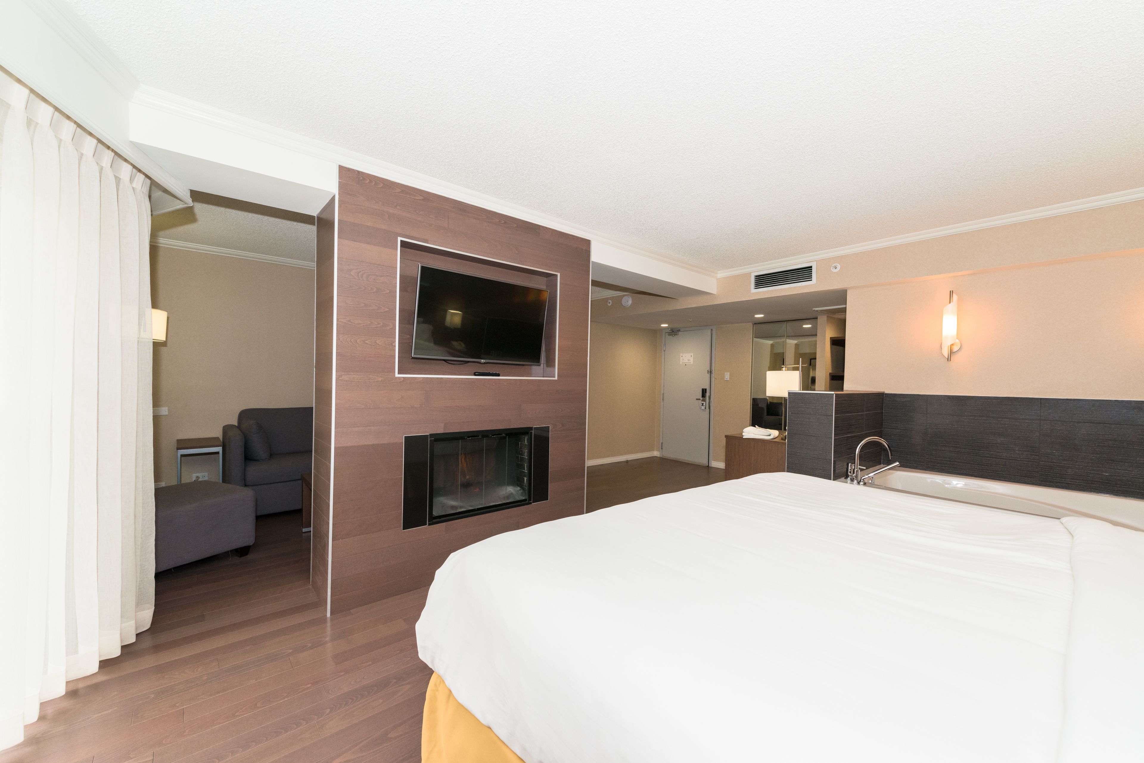 A hotel room with a large bed, a TV mounted on the wall, a small seating area, and a bathroom with a sink and mirror.