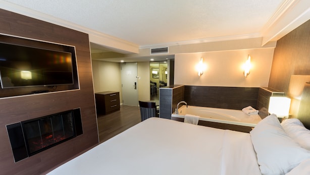 A modern hotel room with a large bed, a TV mounted on the wall, a built-in fireplace, a bathroom with a bathtub, and a walk-in closet.