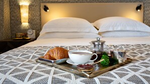 Una cama bien hecha con ropa blanca, una bandeja con croissant, café y mermelada, y una lámpara de pared.