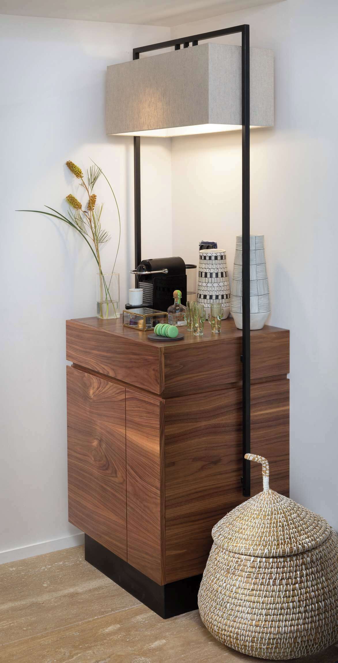 Un escritorio moderno de madera con estructura metálica negra, una lámpara, objetos decorativos y un otoman tejido.