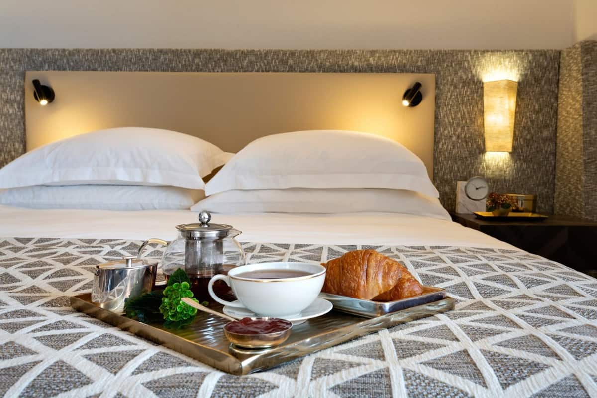 Ein ordentlich bezogenes Bett mit weißen Laken, ein Tablett mit Frühstücksleckereien, darunter Croissant, Tee und Marmelade, sowie ein Nachttisch mit einer Uhr und Dekoration.
