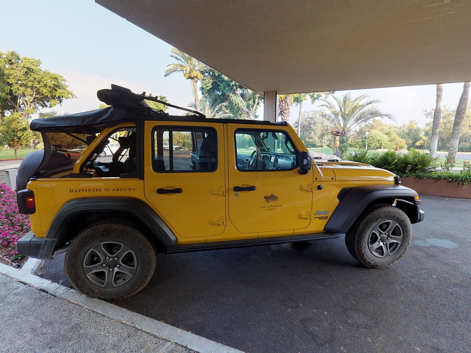 A yellow Jeep Wrangler parked under a covered area with a view of palm trees and greenery.