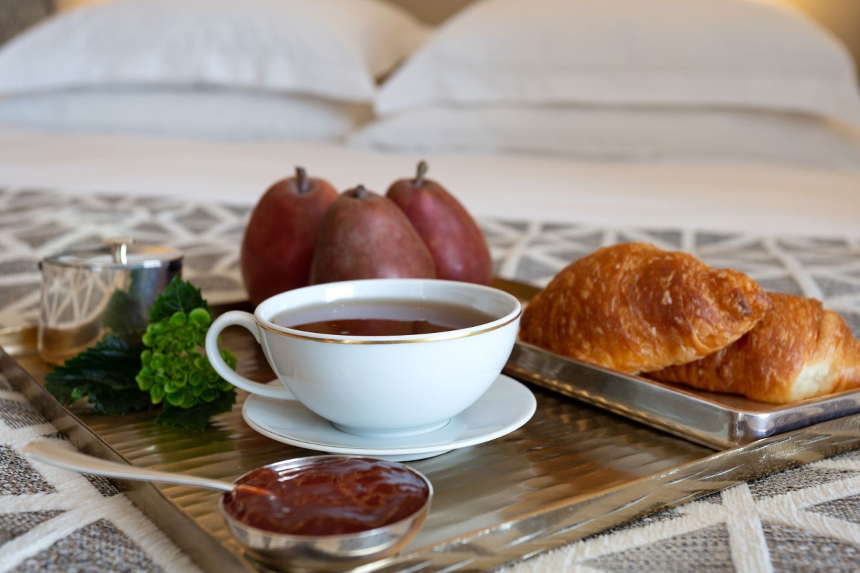 Una bandeja con una taza de té, dos peras, un croissant y un frasco pequeño de mermelada sobre un mantel estampado.