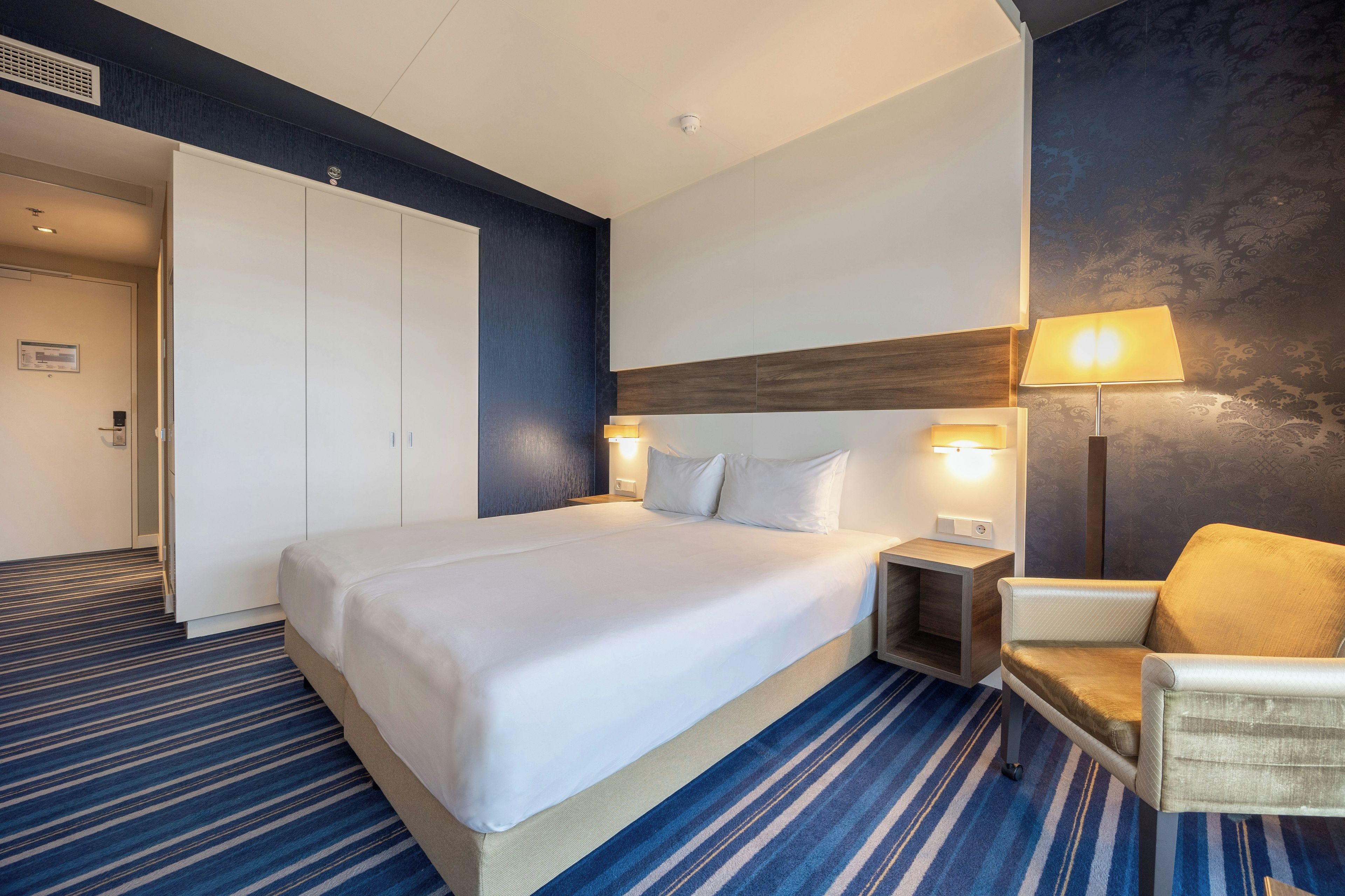 Habitación de hotel con una cama grande, lámparas de noche, una silla y un armario.