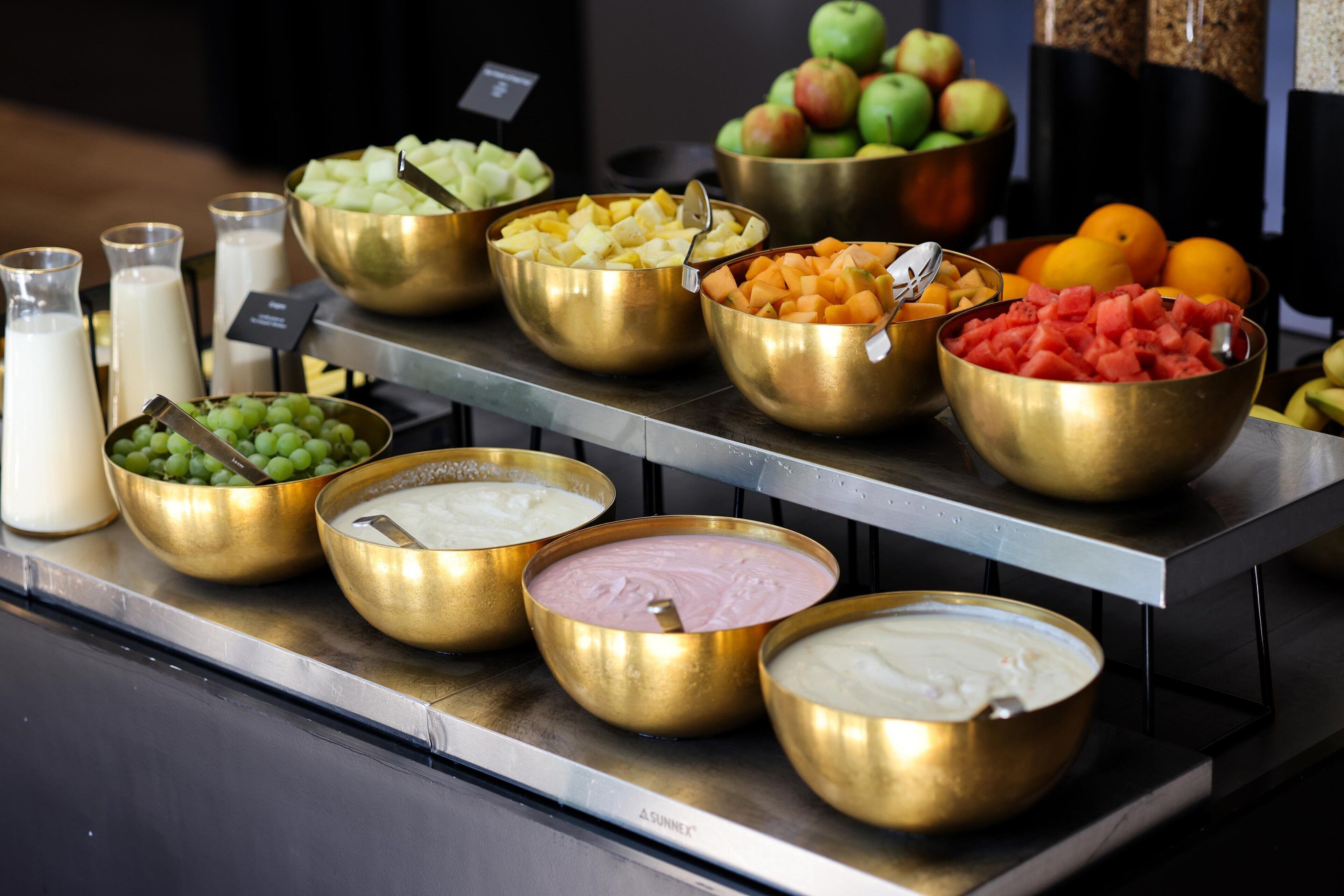 Um buffet com diversos alimentos em recipientes dourados, incluindo frutas e diferentes tipos de molhos.
