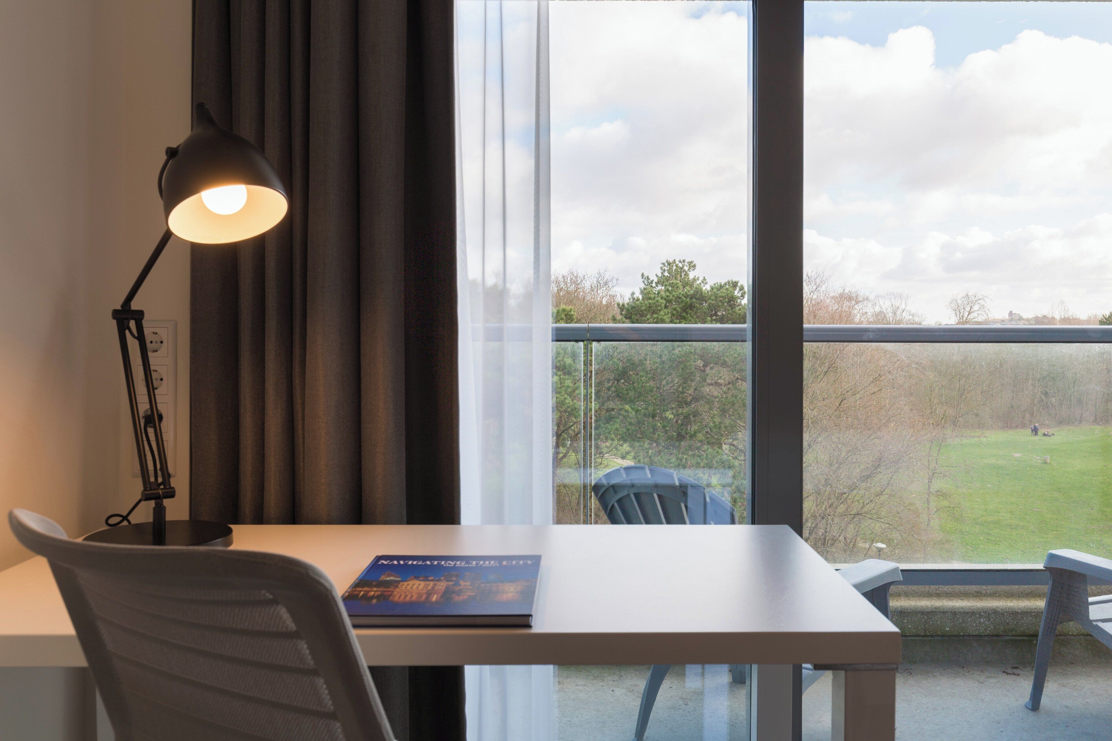 A room with a desk, a chair, a lamp, and a view of a grassy area through a window.