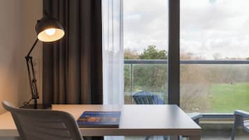 A room with a desk, a chair, a lamp, and a view of a grassy area through a window.