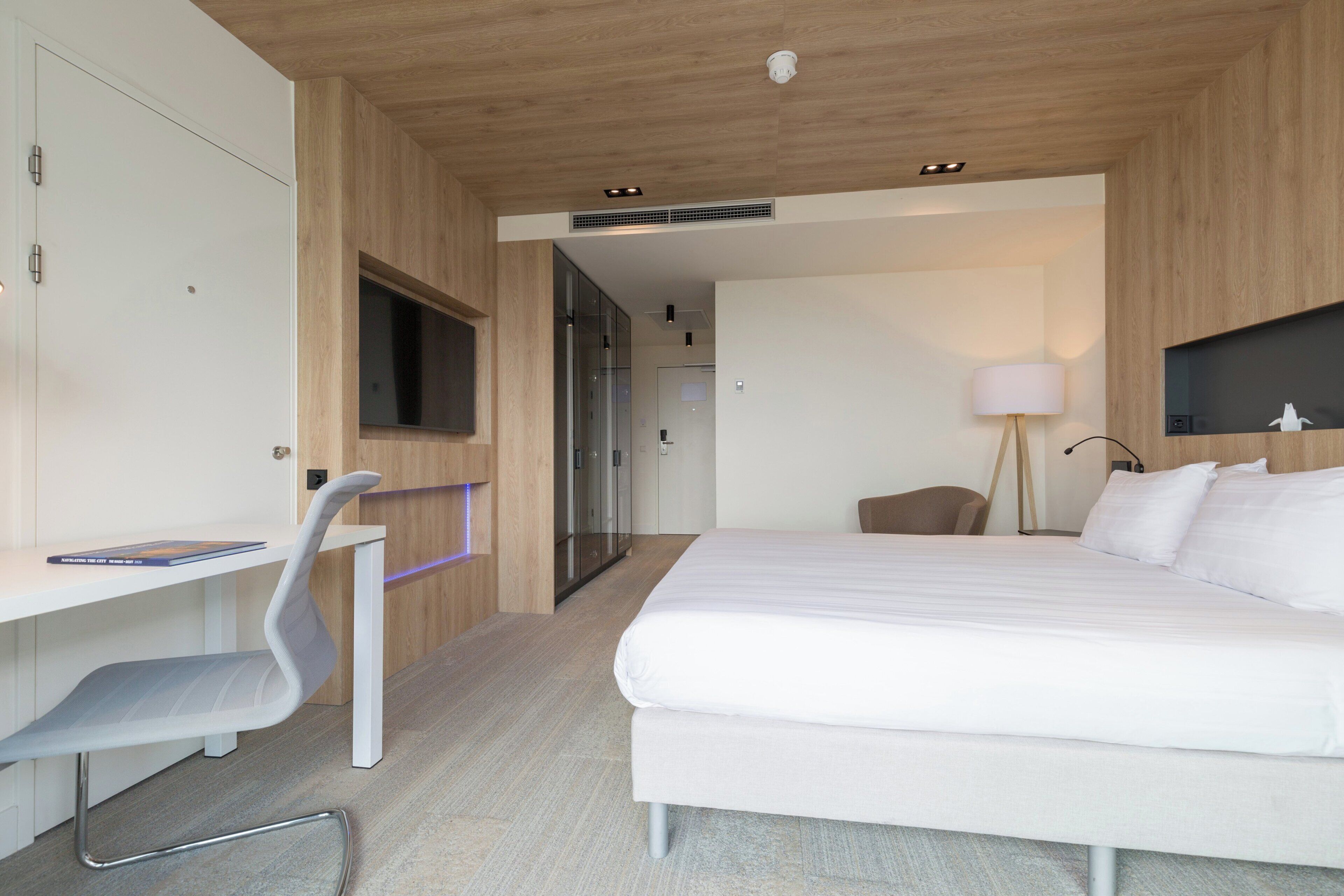 Quarto de hotel moderno com cama, escrivaninha, cadeira e televisão fixada na parede.