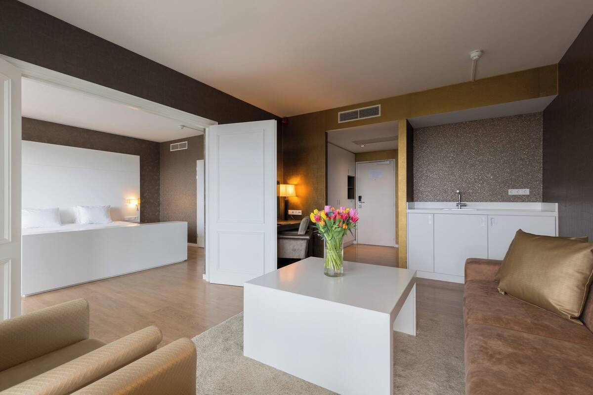 Un salon moderne avec un canapé blanc, un canapé marron, une table basse blanche ornée d’un vase de fleurs, et un coin cuisine en arrière-plan.