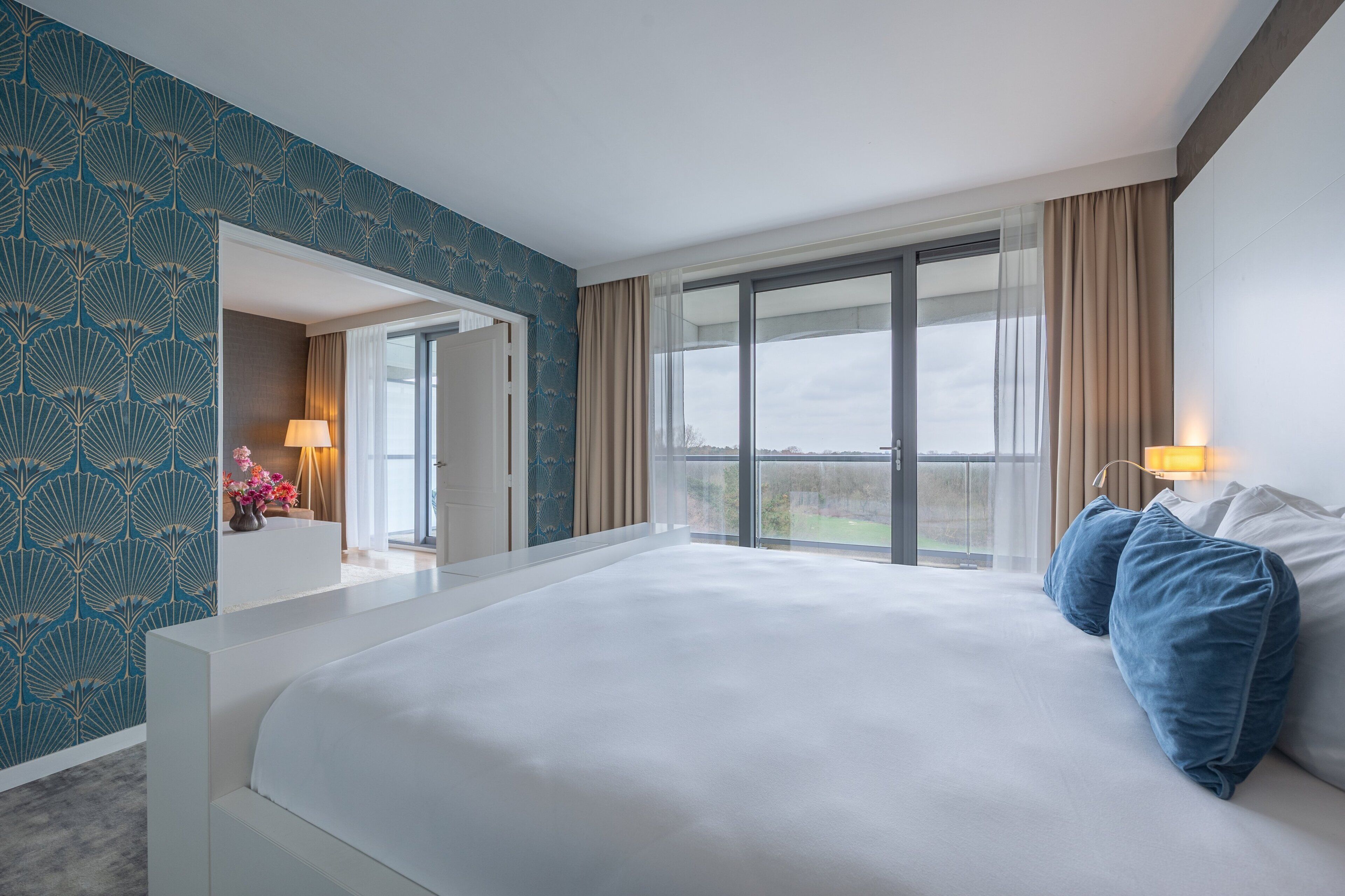 A hotel room with a large bed, a view through a sliding glass door, and a patterned wall.