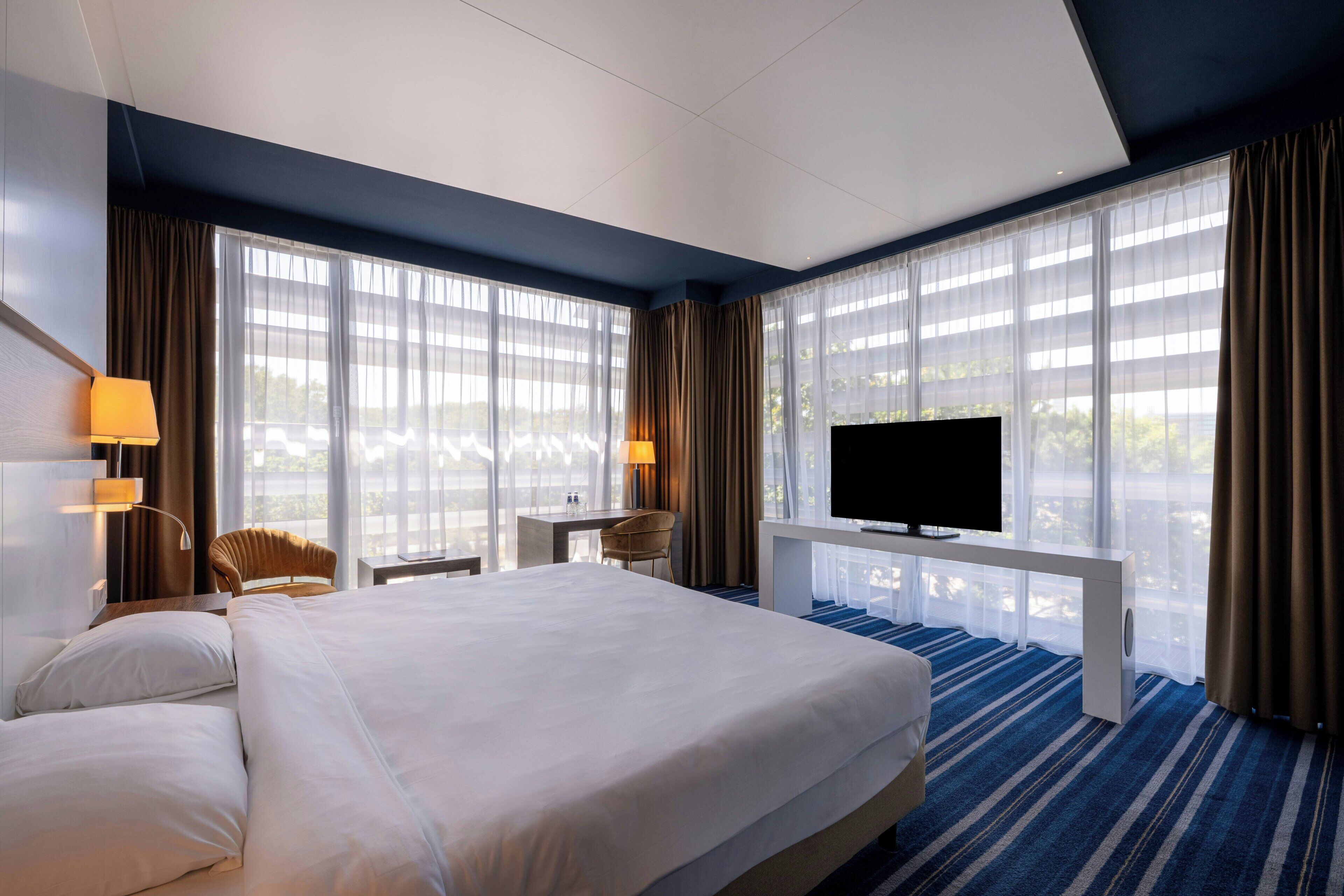Ein Hotelzimmer mit einem großen Bett, einem Fernseher, einem Schreibtisch mit Stuhl und einem fenster mit Vorhängen.