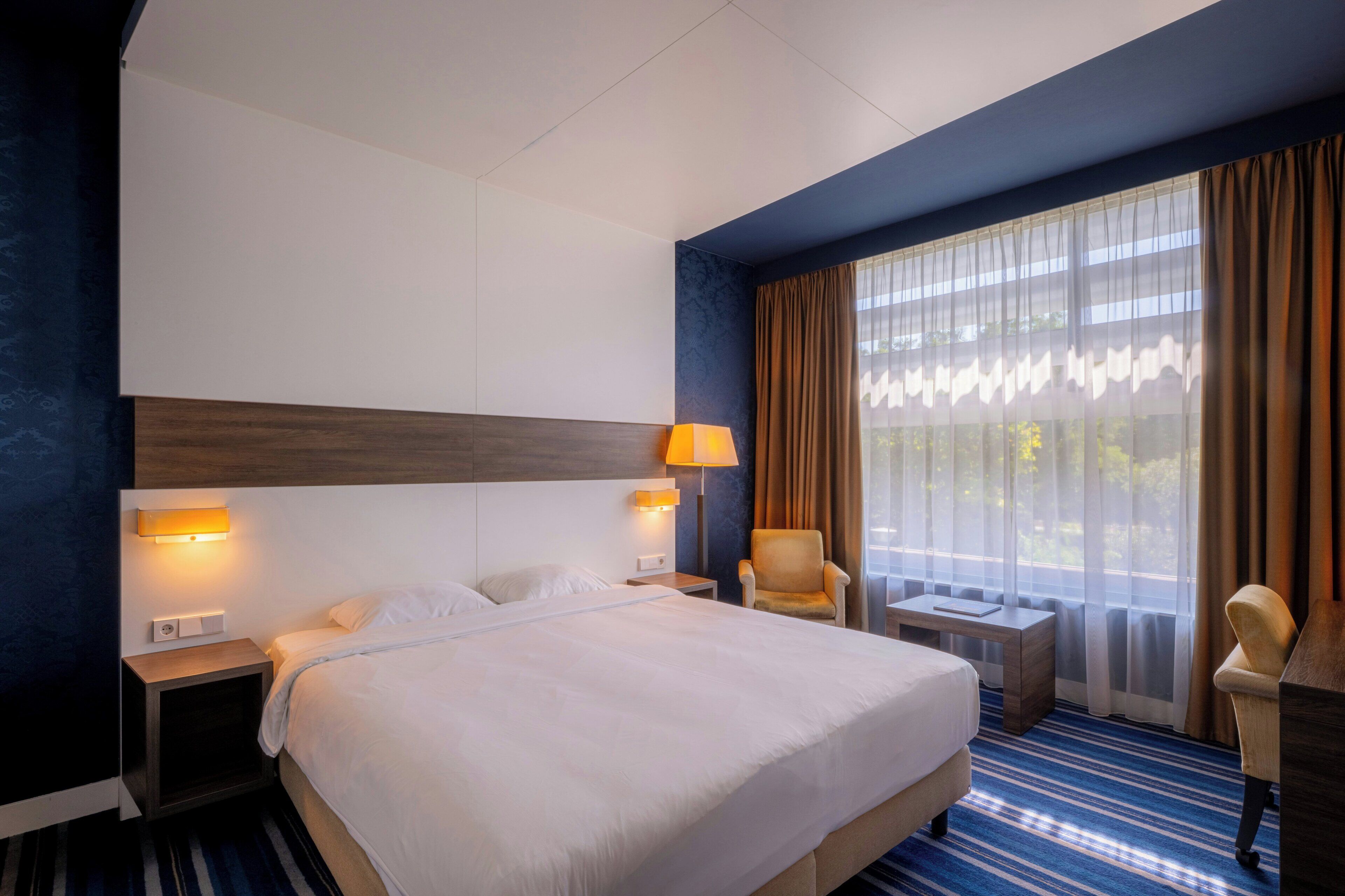 Ein Hotelzimmer mit einem großen Bett, zwei Nachttischen, einem Sessel, einem Schreibtisch und einem Fenster mit Vorhängen.