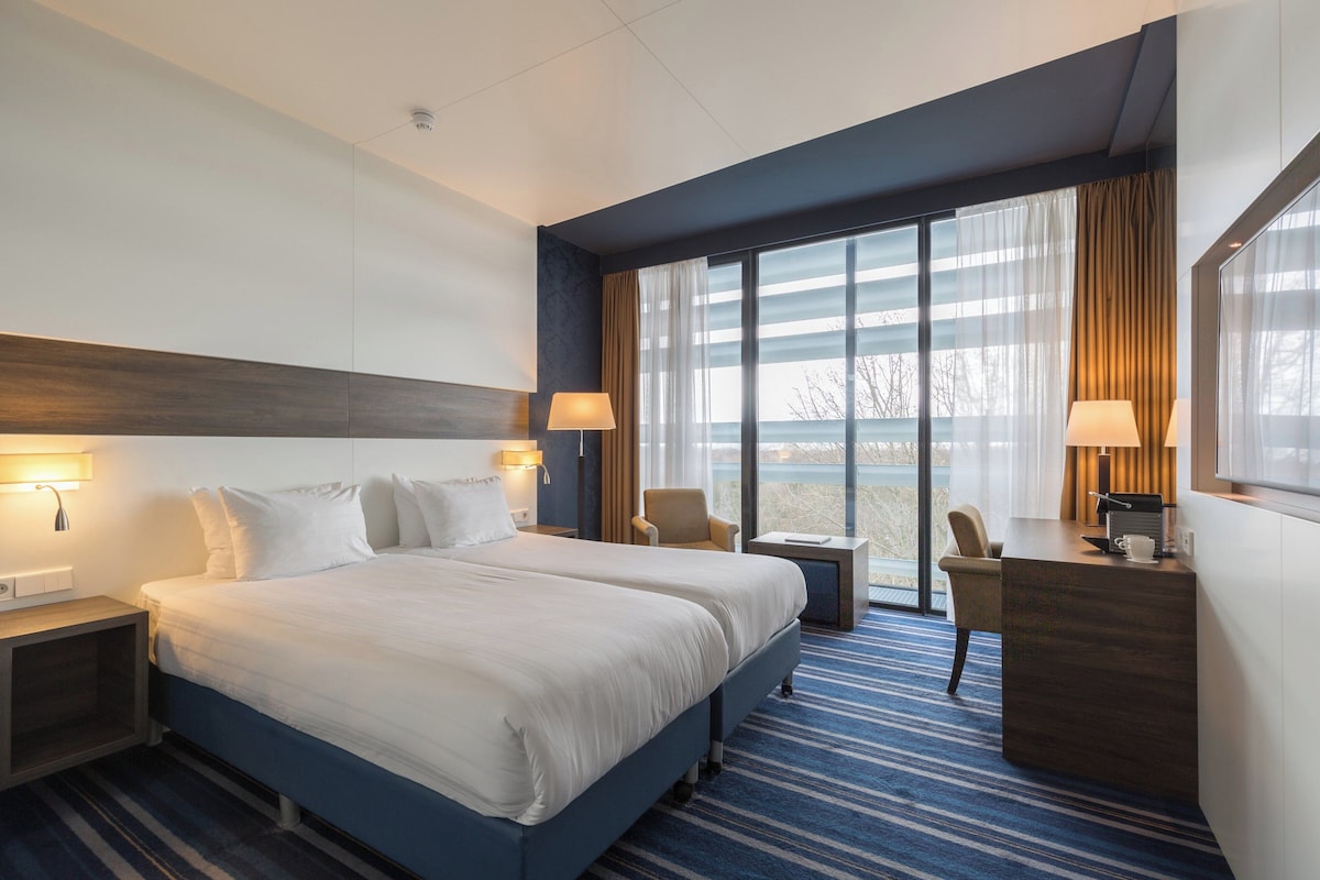 Ein Hotelzimmer mit einem großen Bett, zwei Nachttischen mit Lampen, einem Schreibtisch mit Stuhl und Blick ins Freie durch große Fenster.