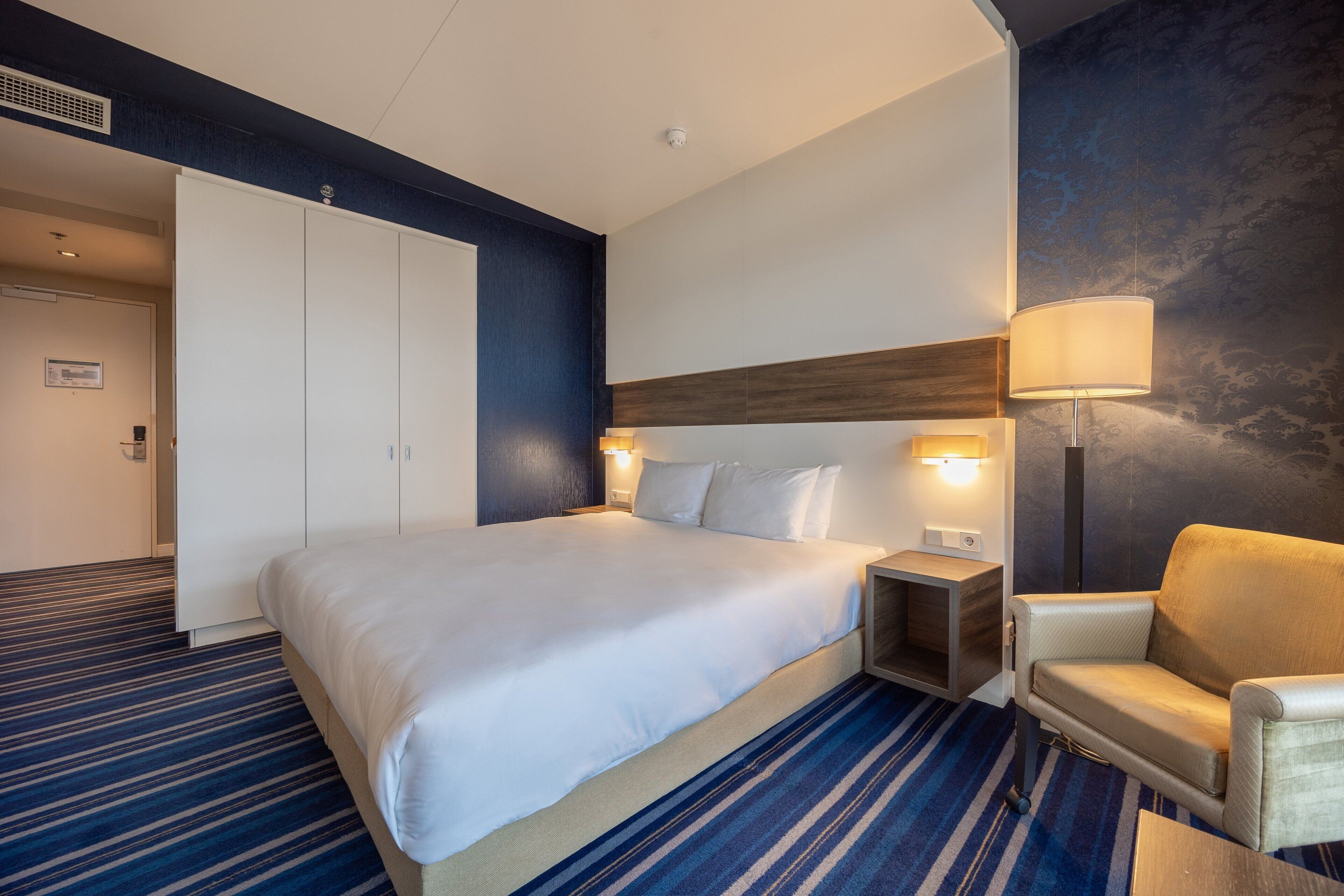 A hotel room with a large bed, a chair, a nightstand, and a lamp.