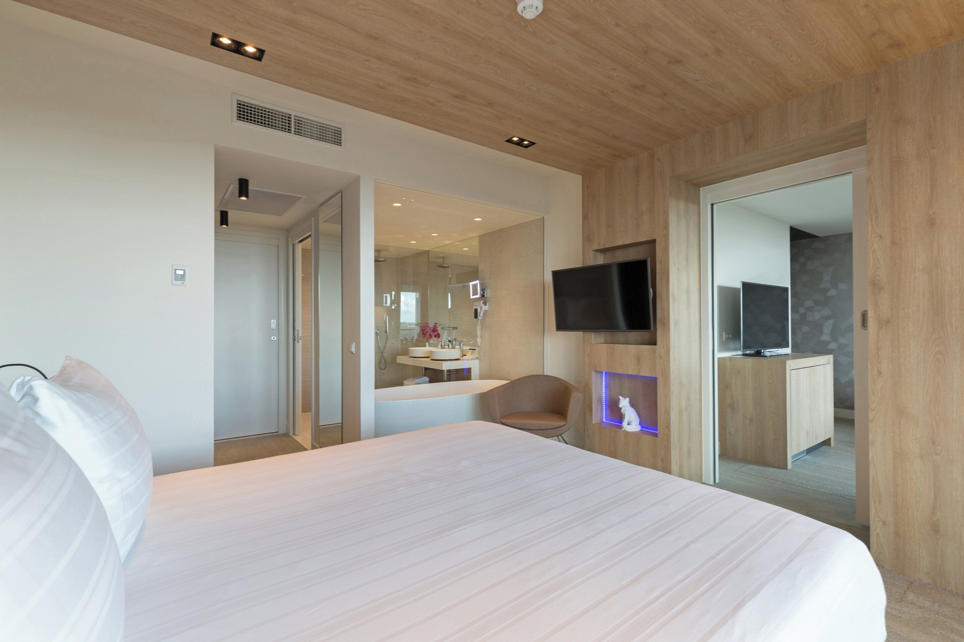 Una habitación de hotel moderna con una cama grande, un televisor de pantalla plana montado en la pared y una pequeña zona de estar con una silla.