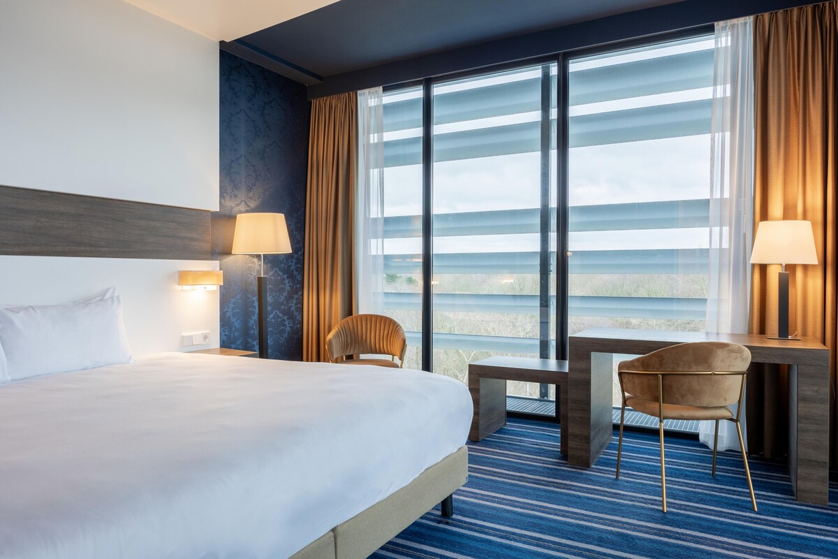 A hotel room with a large bed, two chairs, a desk, and a view through large windows.