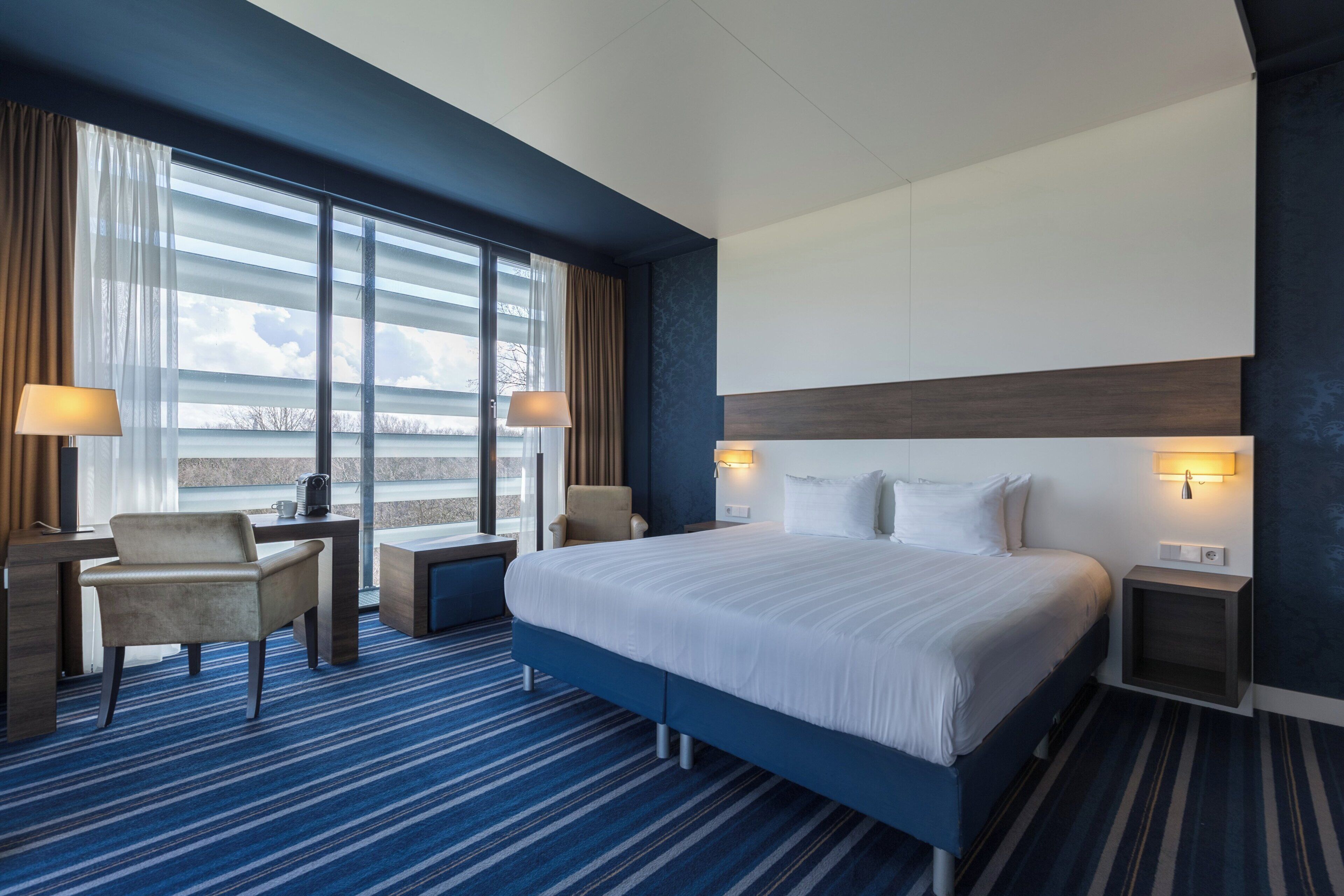 Une chambre d’hôtel avec un grand lit, deux tables de chevet, un bureau avec une chaise et une vue sur l’extérieur grâce à de grandes fenêtres.