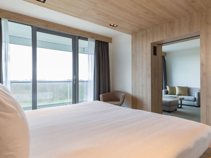 Een moderne hotelkamer met een groot bed, een stoel, een bank en uitzicht op het omliggende landschap door grote ramen.