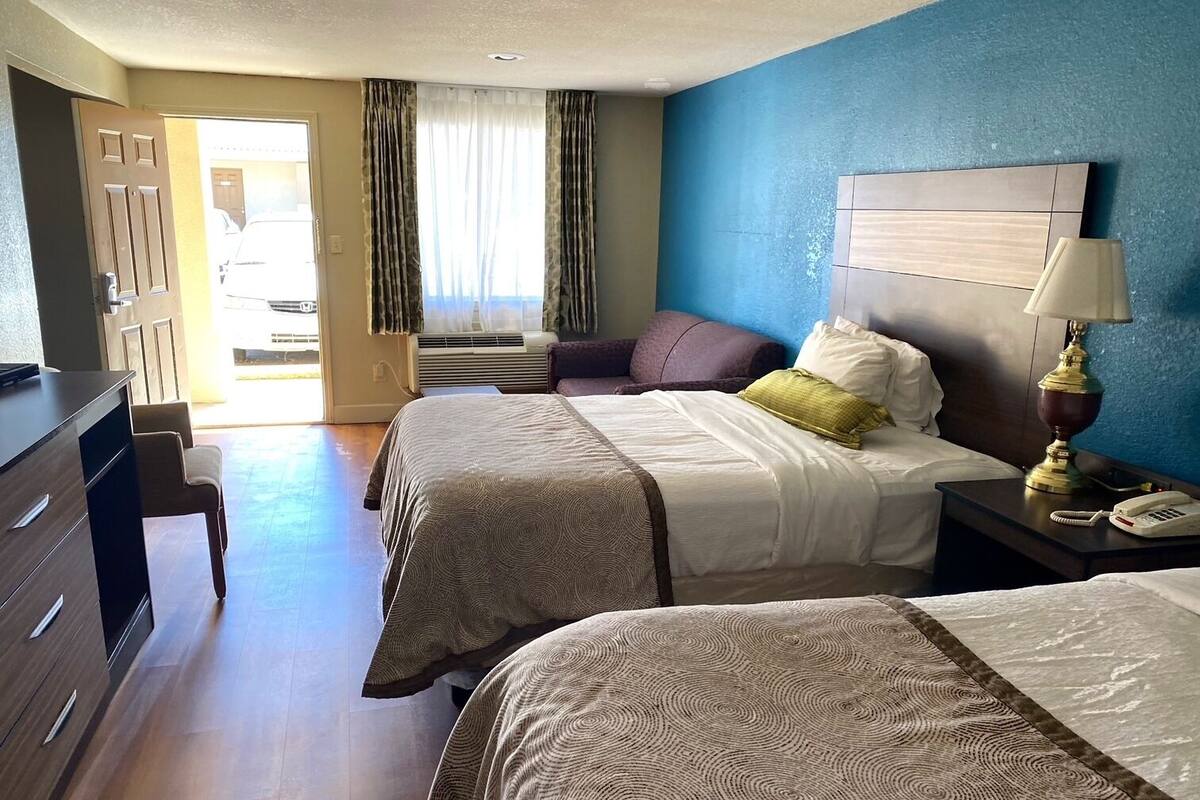 A hotel room with two beds, a desk, a chair, a lamp, and a view of the outside.