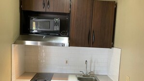 A compact kitchen with dark wooden cabinets, a built-in microwave, a stainless steel sink, and a white tiled backsplash. - Stratford House Inn and Suites (Temple)