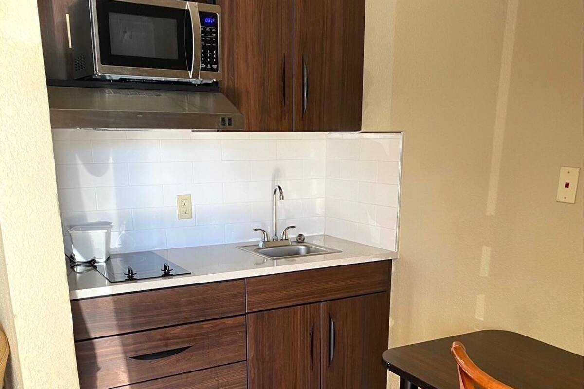 A compact kitchen with dark wooden cabinets, a stainless steel microwave, a sink, and a small dining table with a chair.