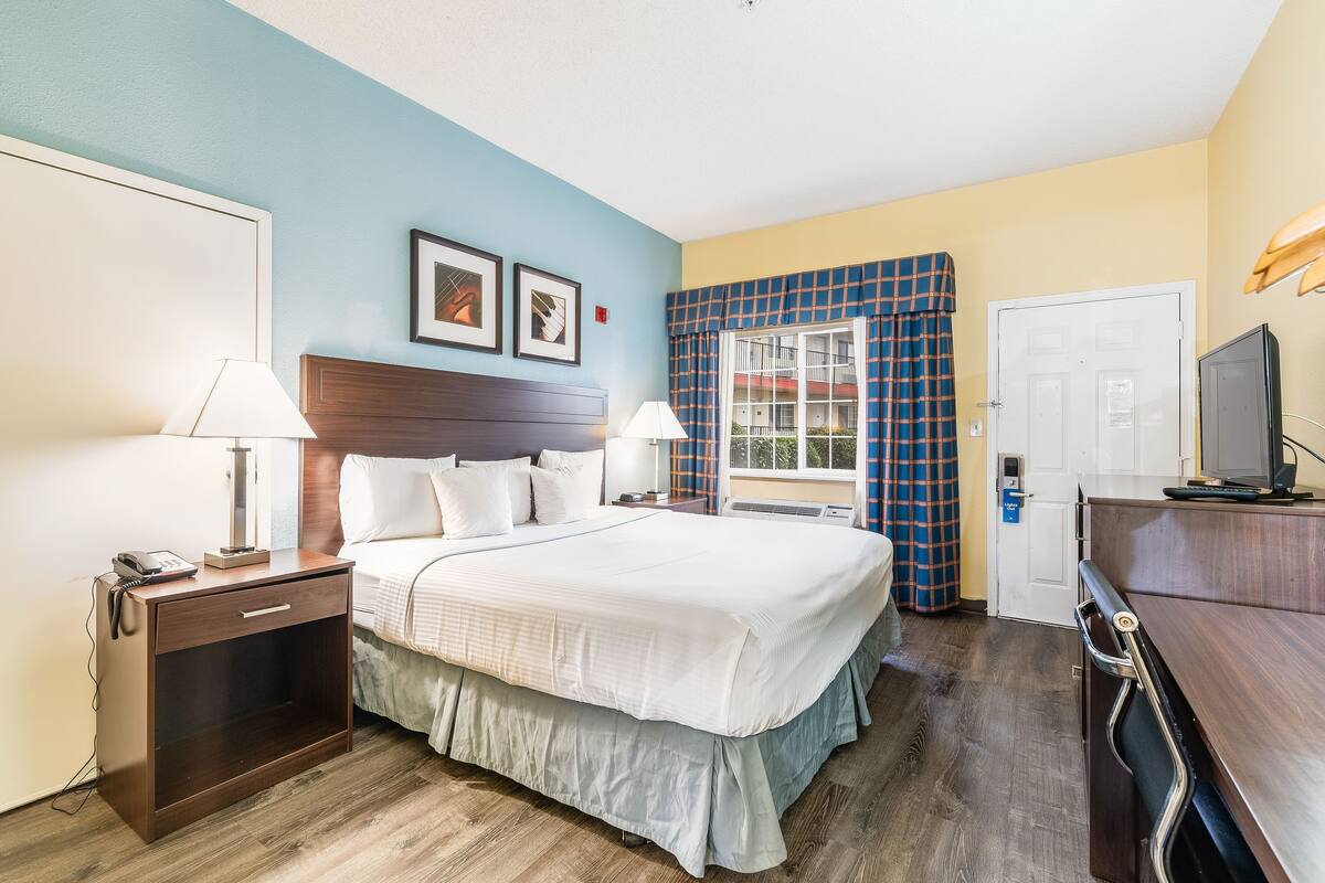 A hotel room with a bed, bedside tables, a desk, a television, and a window with curtains.