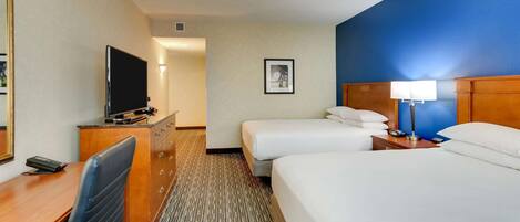 Habitación de hotel con escritorio, dos camas, televisor, lámpara y un cuadro en la pared.